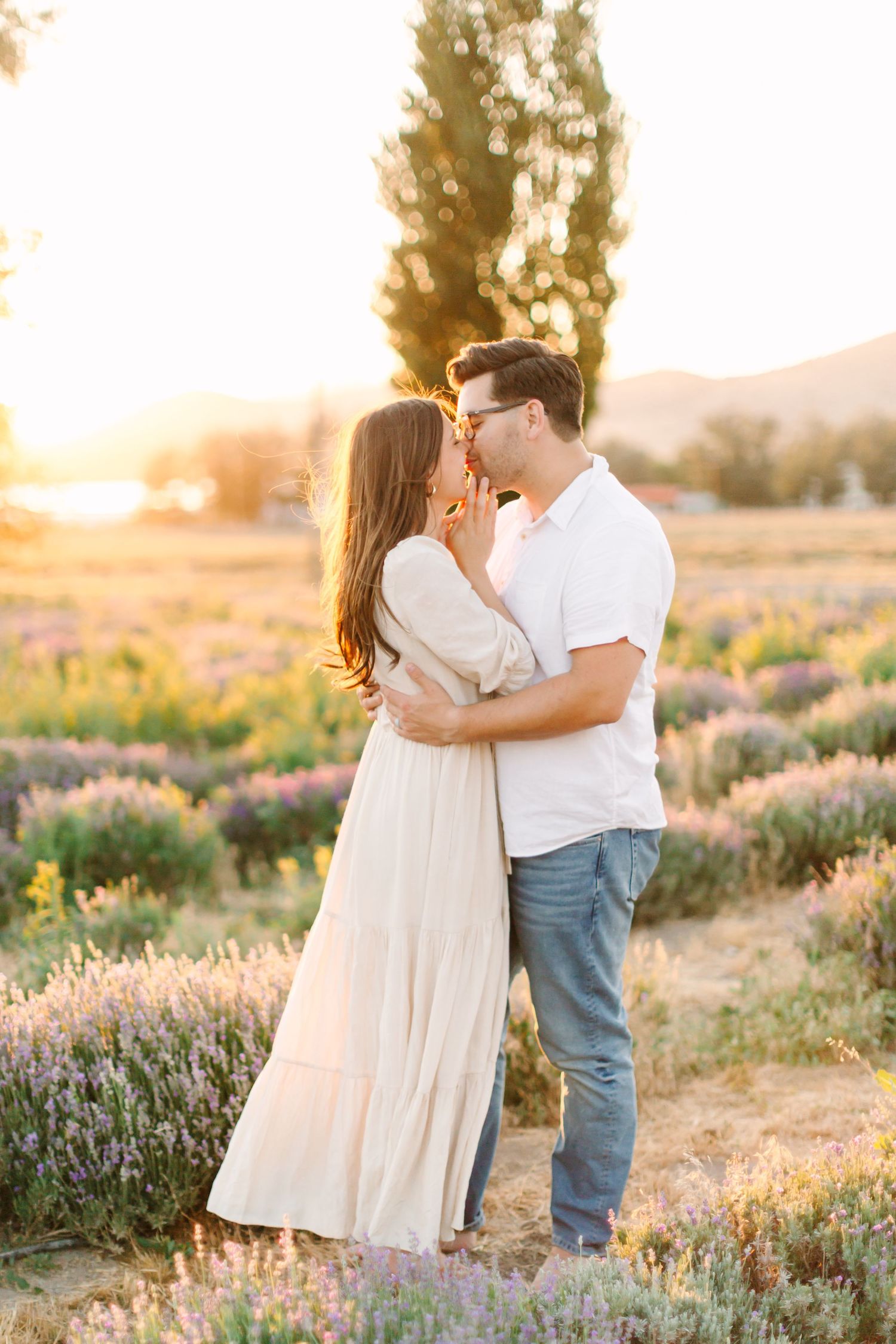A romantic couple embraces in a lavender field at sunset with golden light filtering through trees in the background.