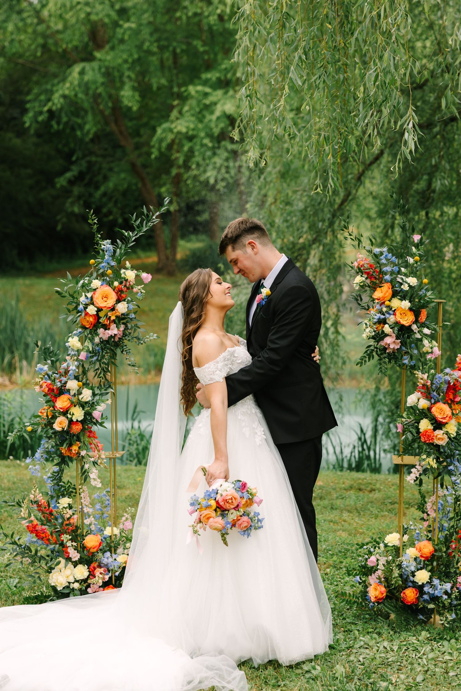 Wedding couple poses near a lake with vibrant orange and pink floral arch arrangements in an outdoor garden setting.