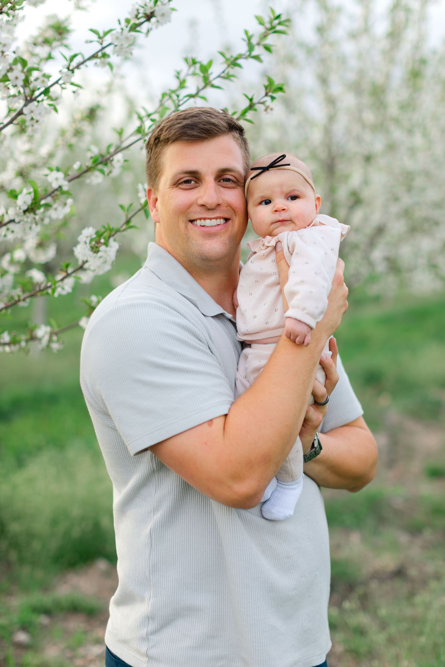 A parent and baby pose together in a blooming spring orchard with white blossoms in the background.