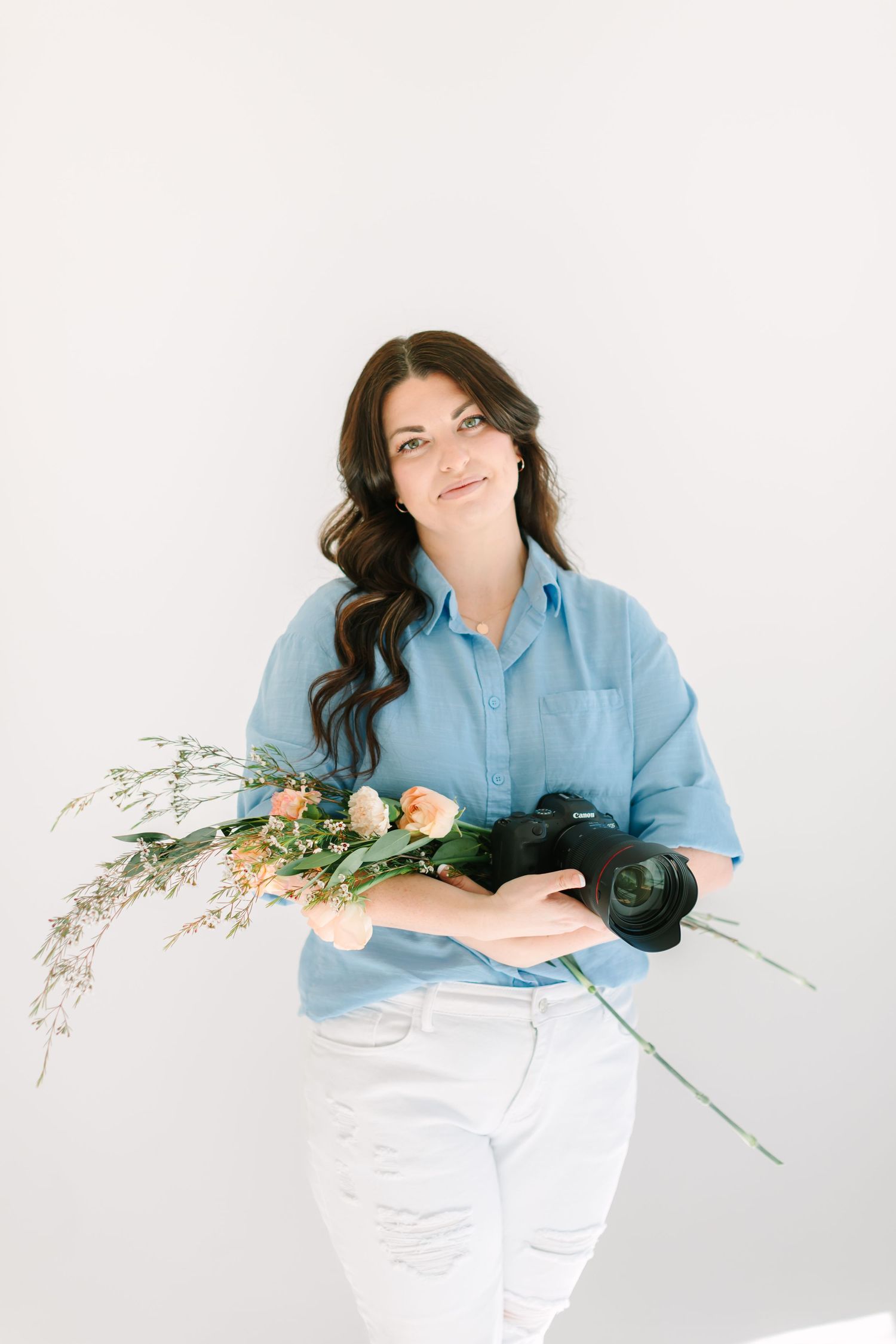 Photographer with camera poses playfully with flowers against minimalist white backdrop.