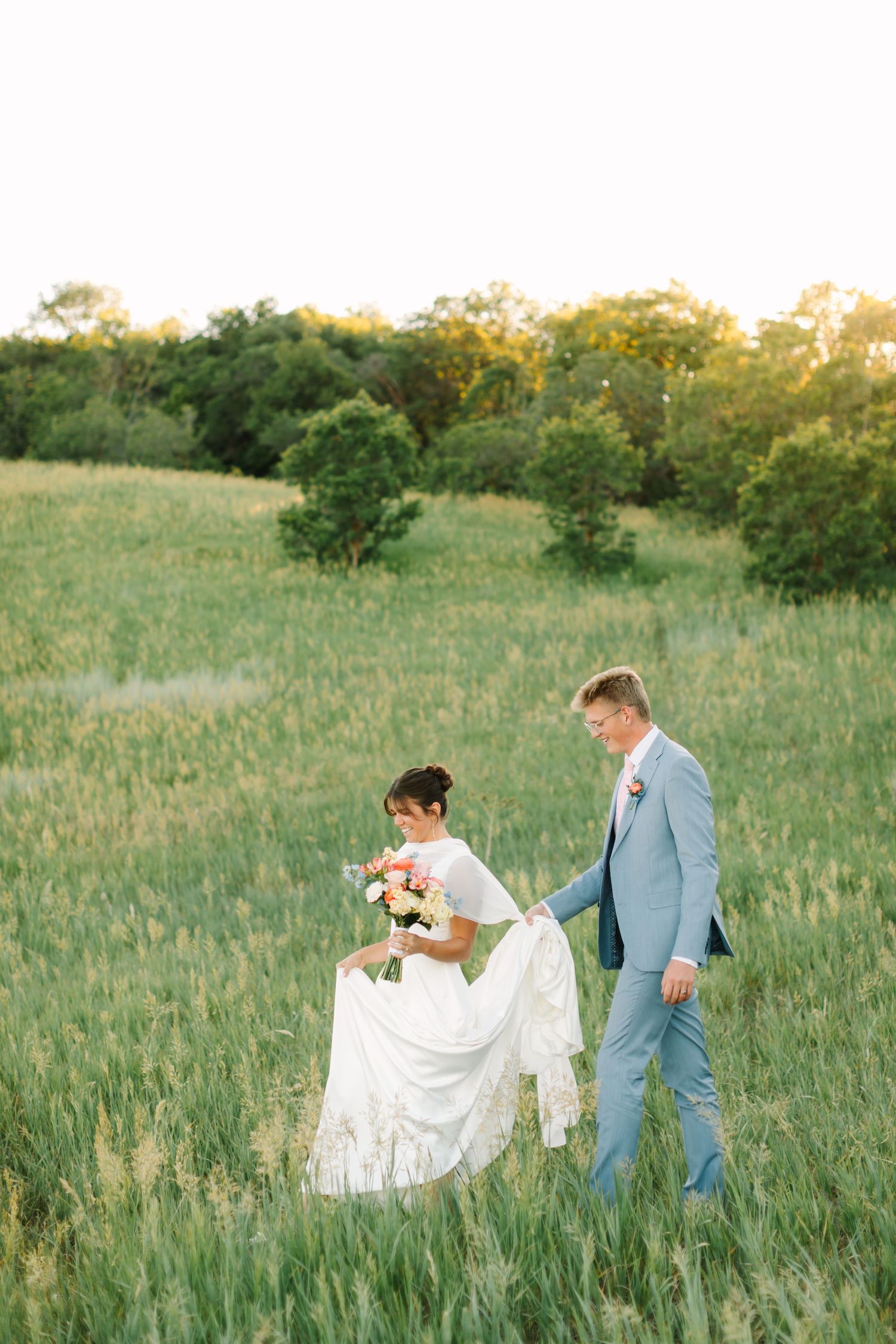 Bride and groom walk through lush green field at golden hour, groom holding bride's white lace dress train.