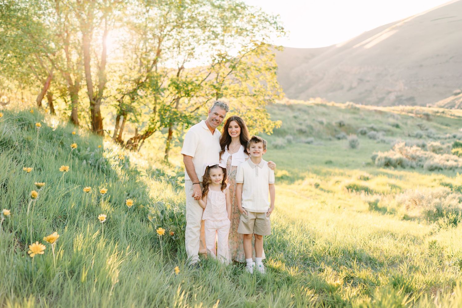 Family of four posing in a sunny green meadow with yellow wildflowers and rolling hills in the background.