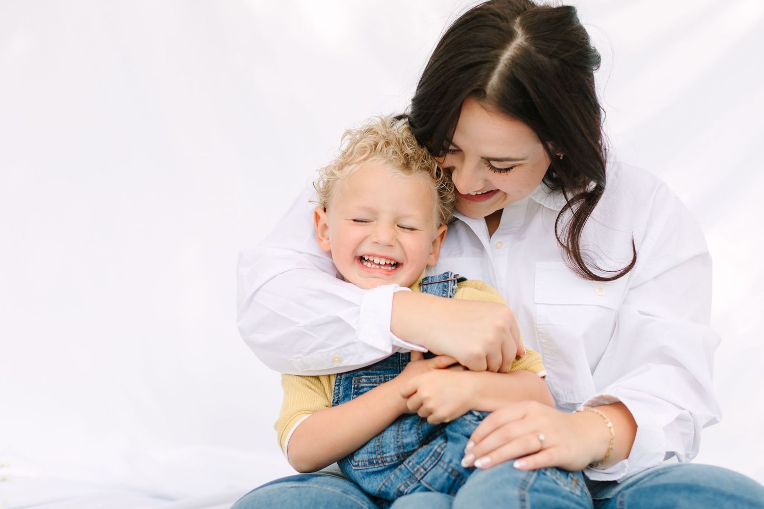 A joyful mother in a white shirt hugs her laughing blond toddler son against a white background.