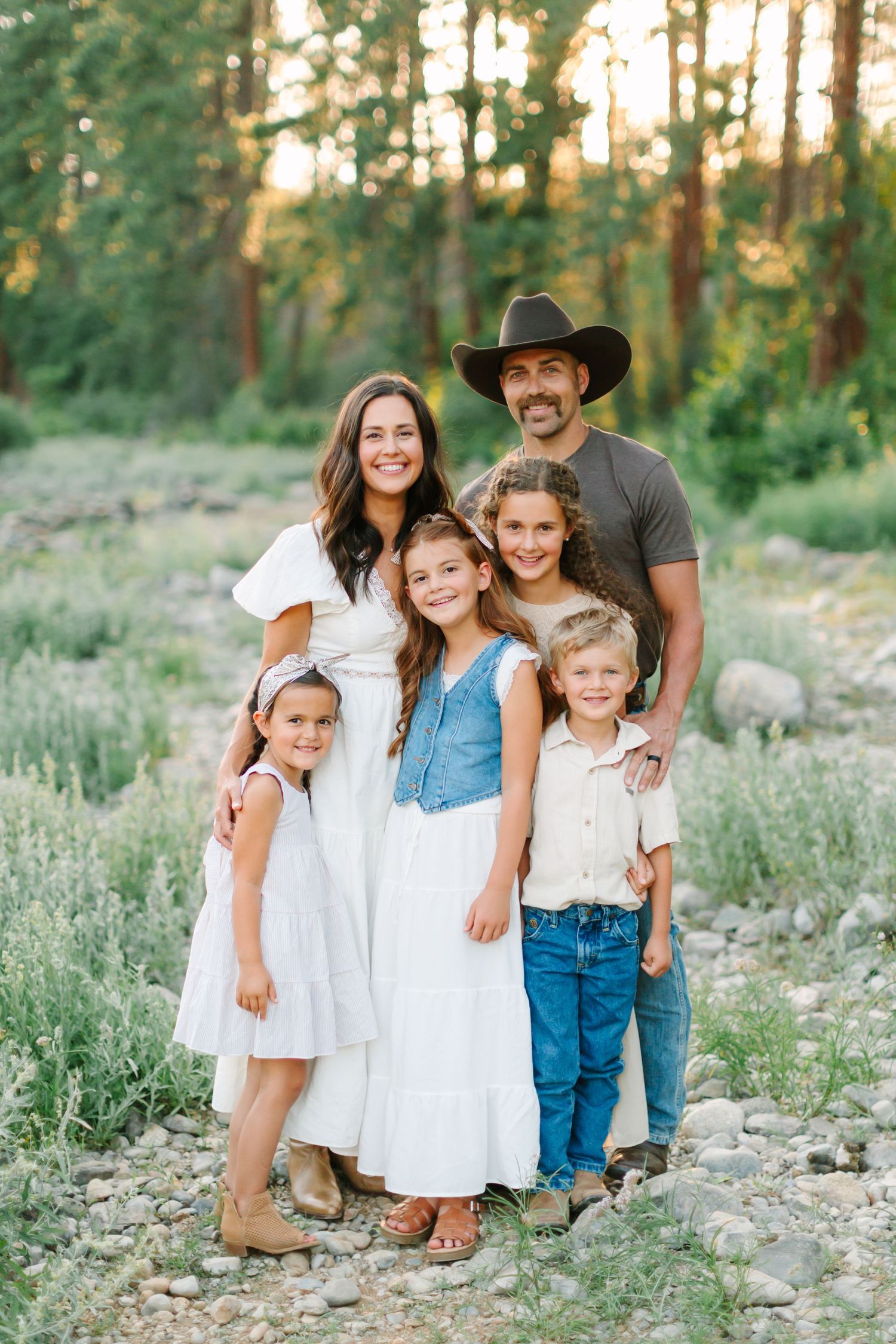 Family in coordinated white and blue outfits poses together in a sunlit forest setting during golden hour.