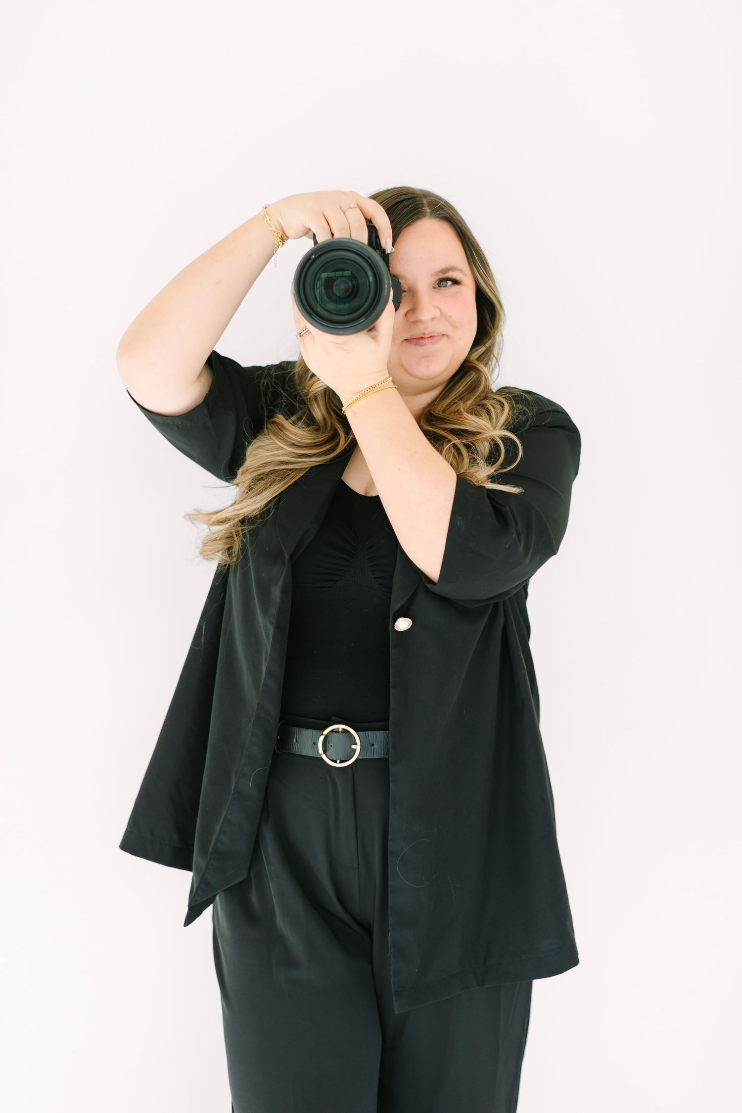 A photographer in all black attire holds up a professional camera against a white background.