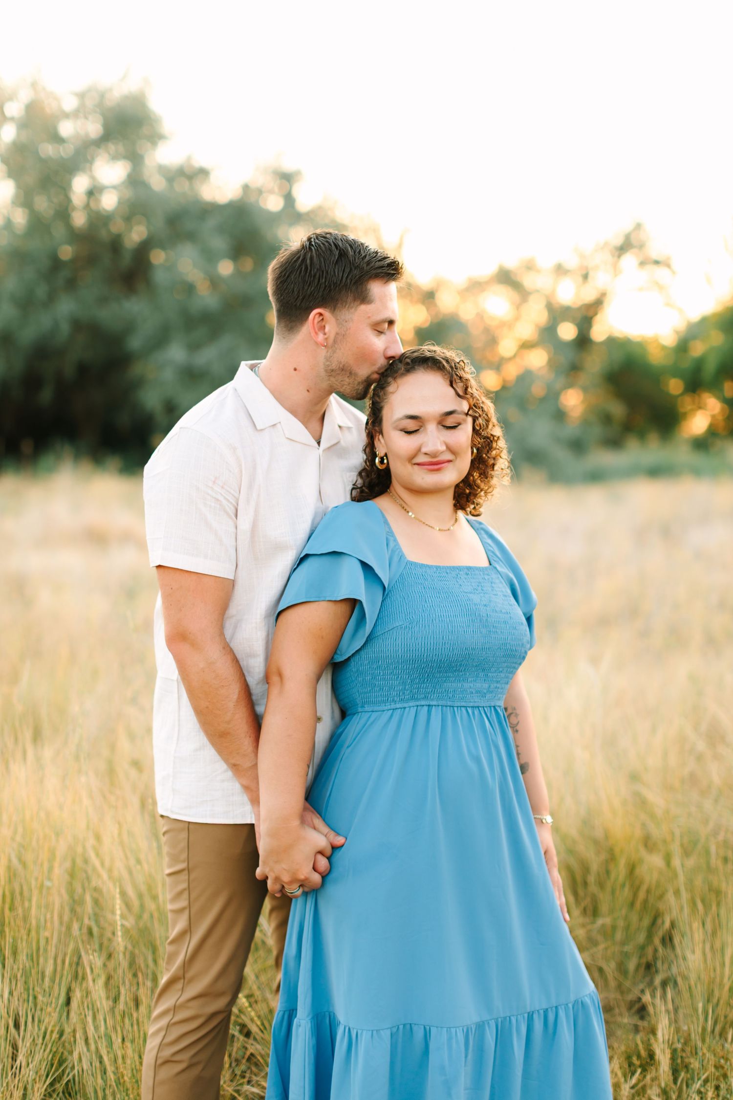 Couple embracing in golden field at sunset, woman in blue dress holding hands with man in white shirt in Bountiful, Utah.