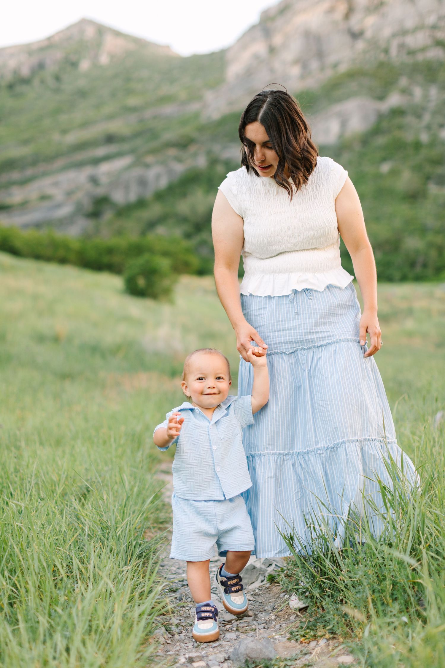 Mother and toddler son walk hand-in-hand on a mountain trail, both dressed in matching light blue outfits.