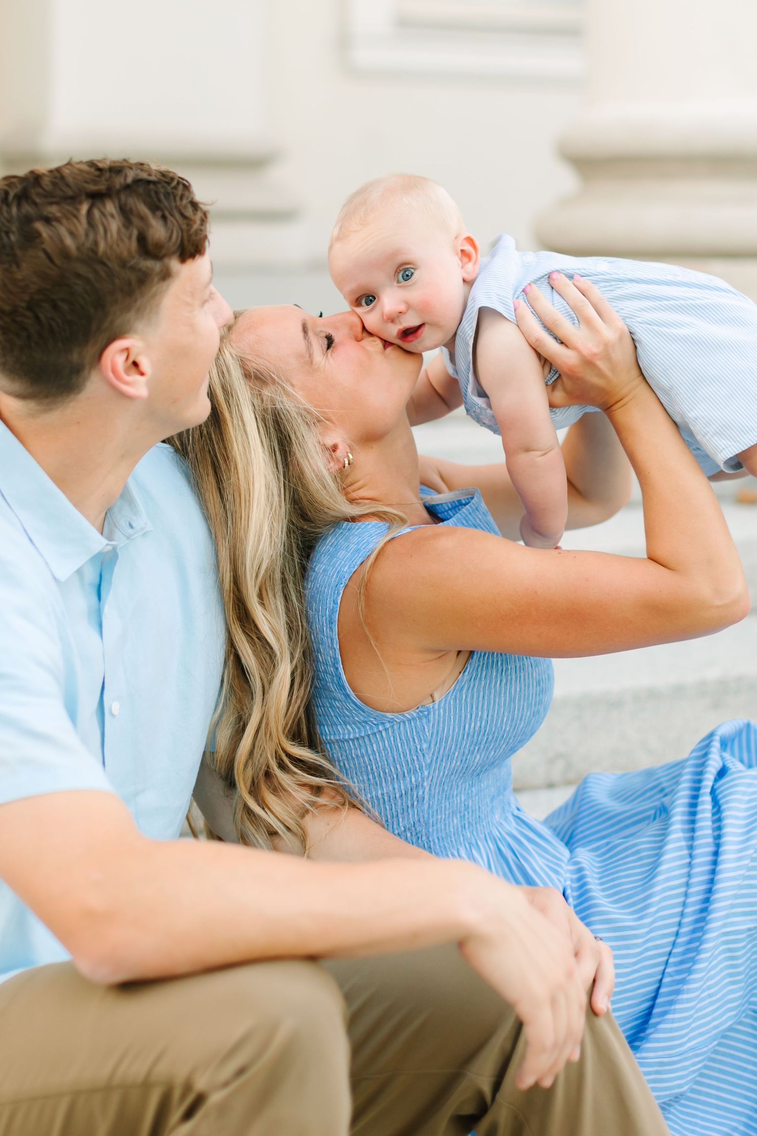 A young family sits on steps as a blonde mother kisses her blue-eyed baby while the father looks on lovingly.