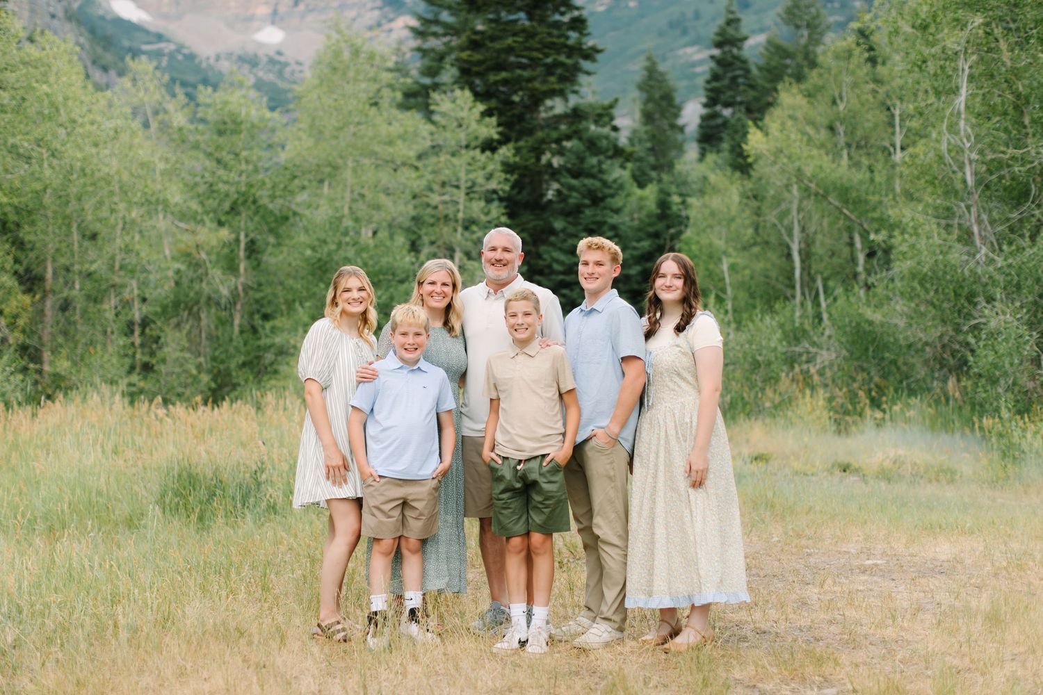 Large family group smiling outdoors in coordinated sage green and white outfits with lush green trees behind in Provo, Utah.