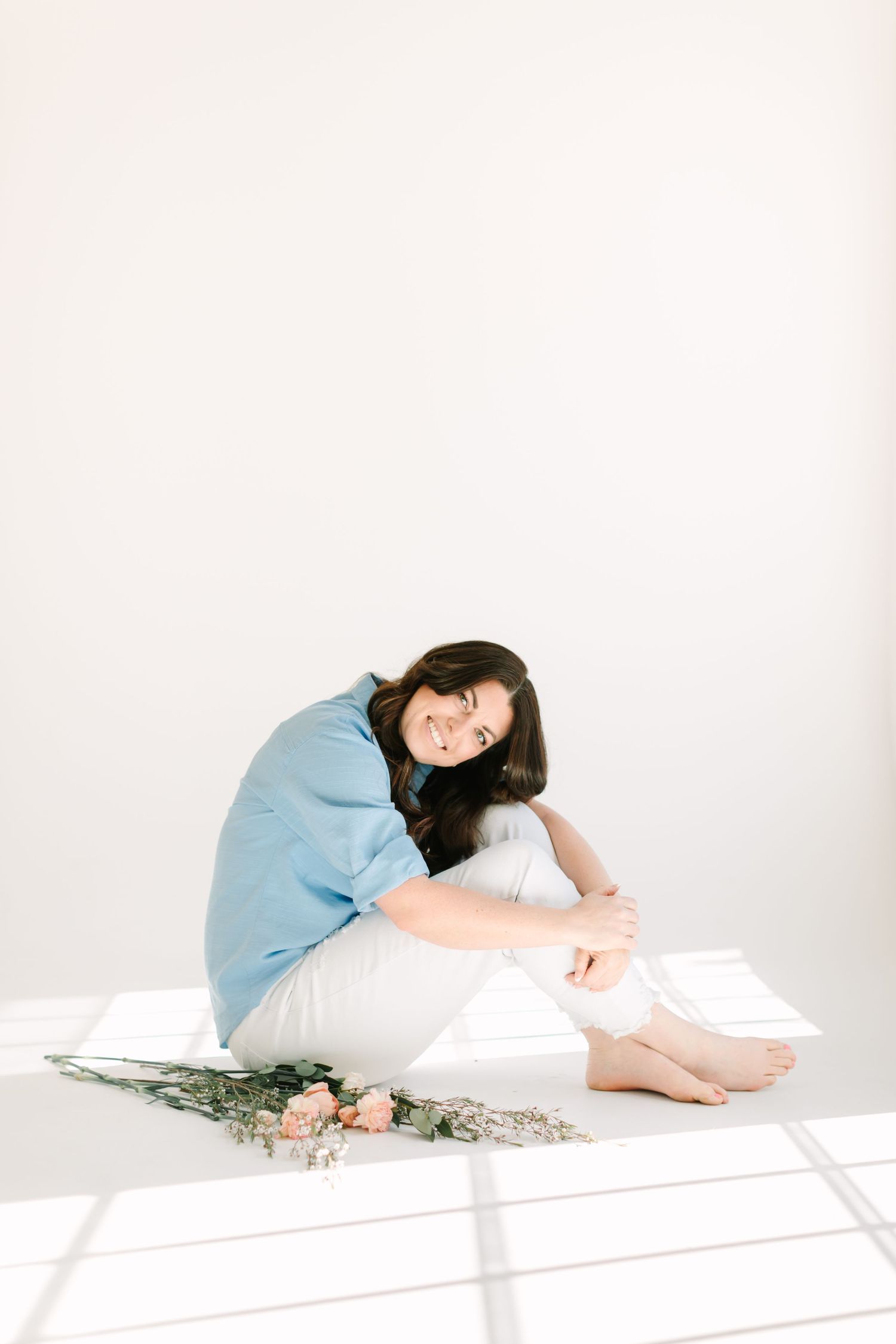 A person in a light blue shirt poses artistically on a white floor with scattered flowers.