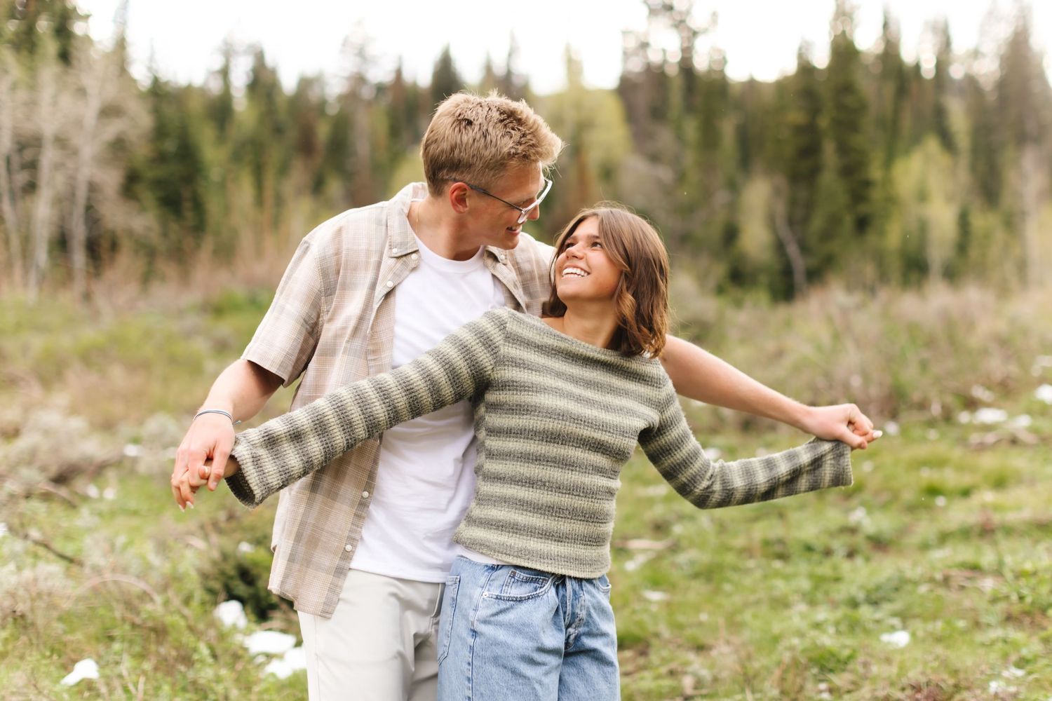 A joyful couple embraces and laughs together in a lush green meadow surrounded by pine trees.