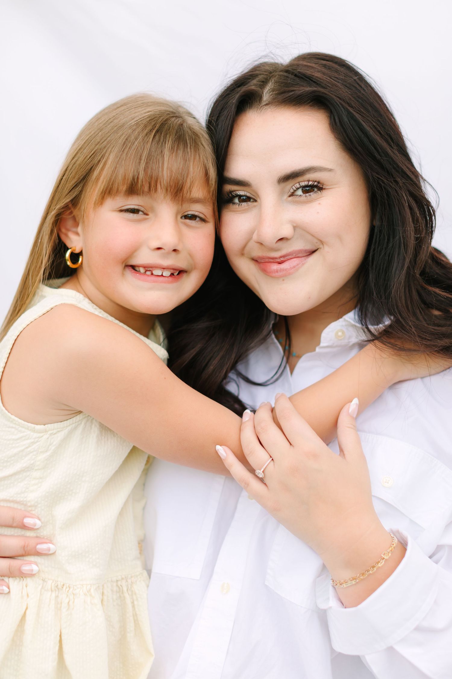 A young girl in a yellow dress hugs a dark-haired woman in a white shirt, both smiling warmly against a white background.