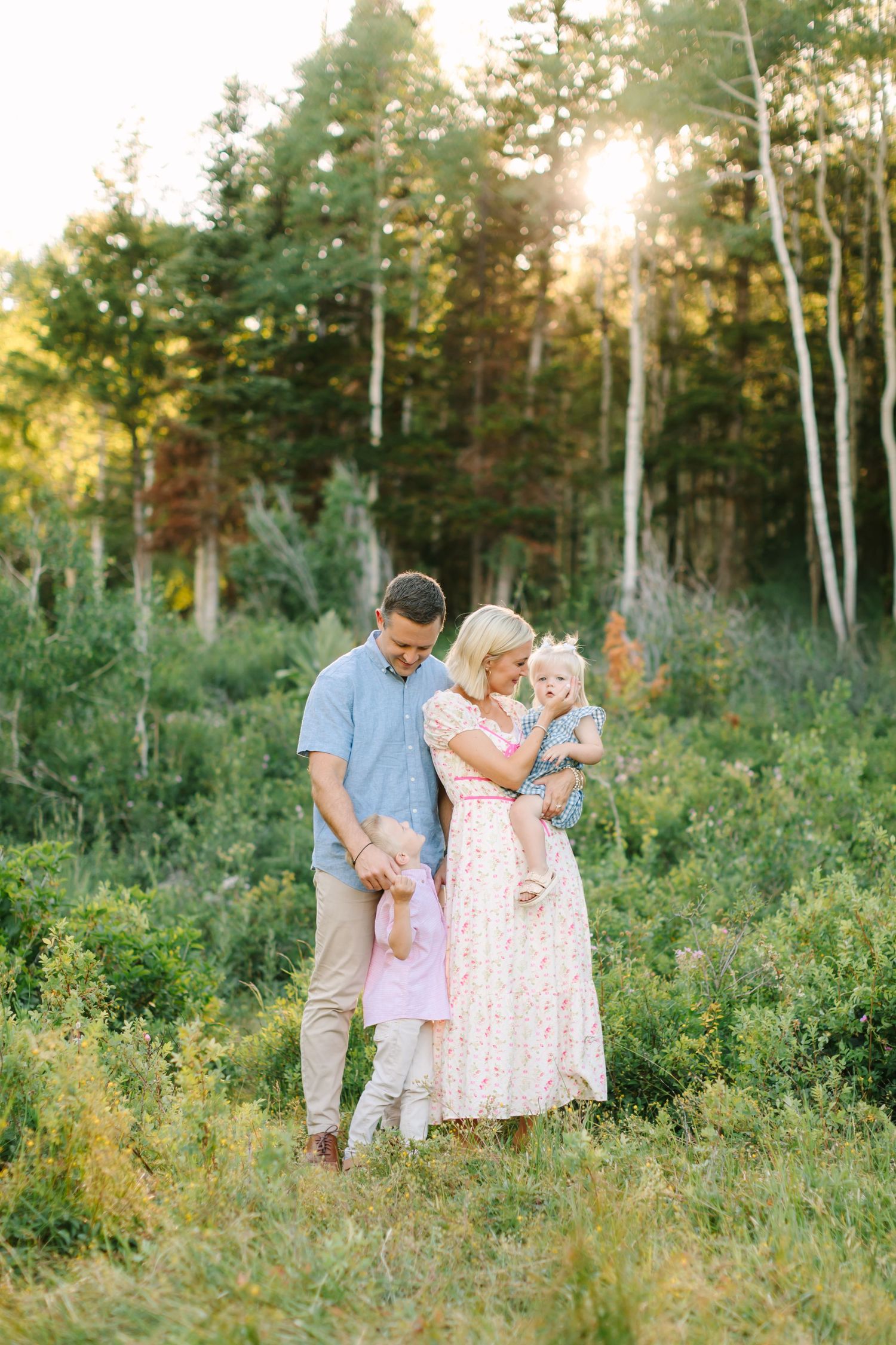 Family of three posing in a sunlit forest meadow, mother in floral dress holding baby, father in blue shirt beside them.