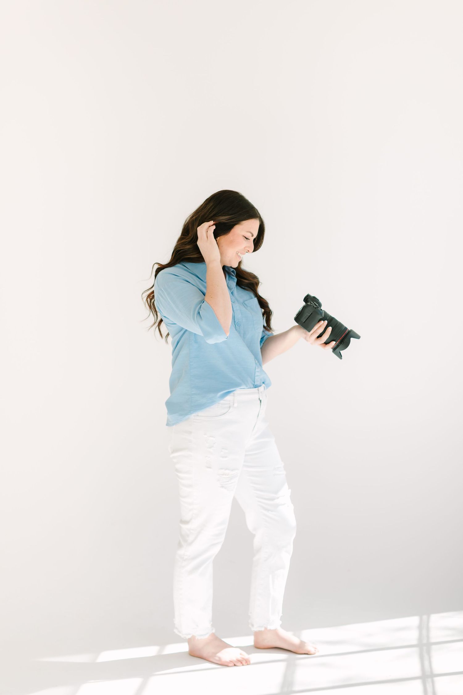 Professional photographer in light blue shirt and white pants holds camera while standing against white wall.