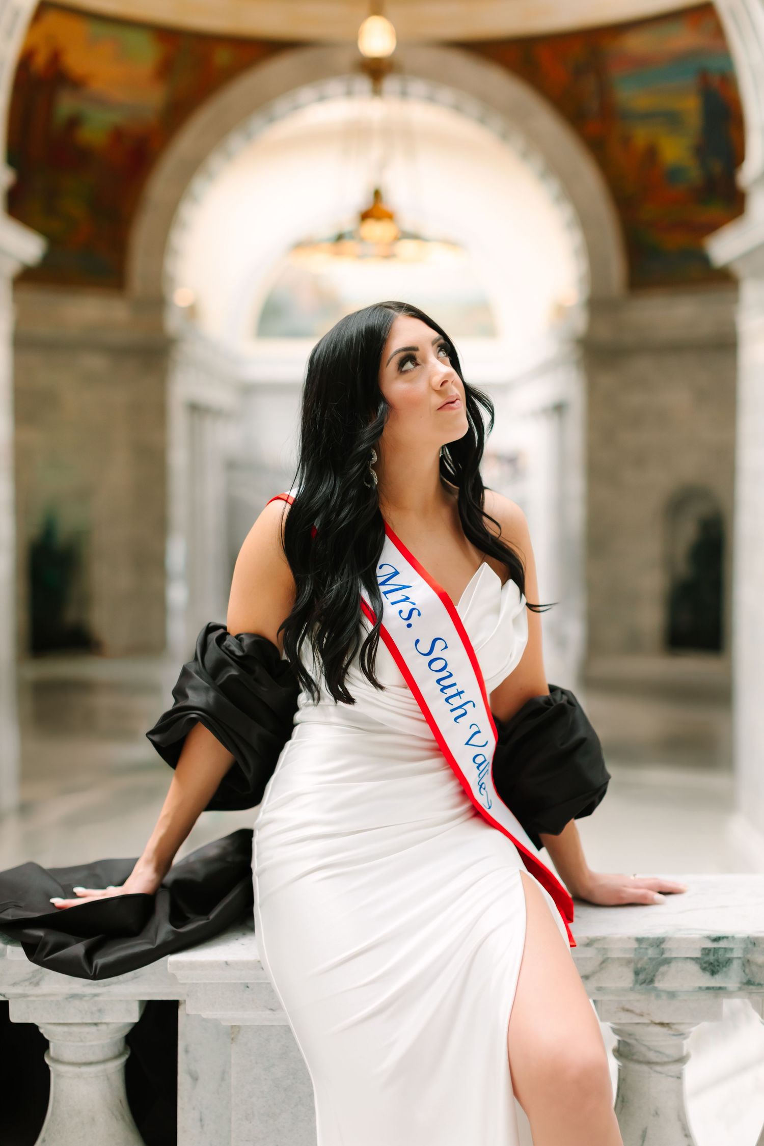 Woman in white dress and black jacket poses elegantly in ornate columned hallway with arched ceilings and pendant lights.