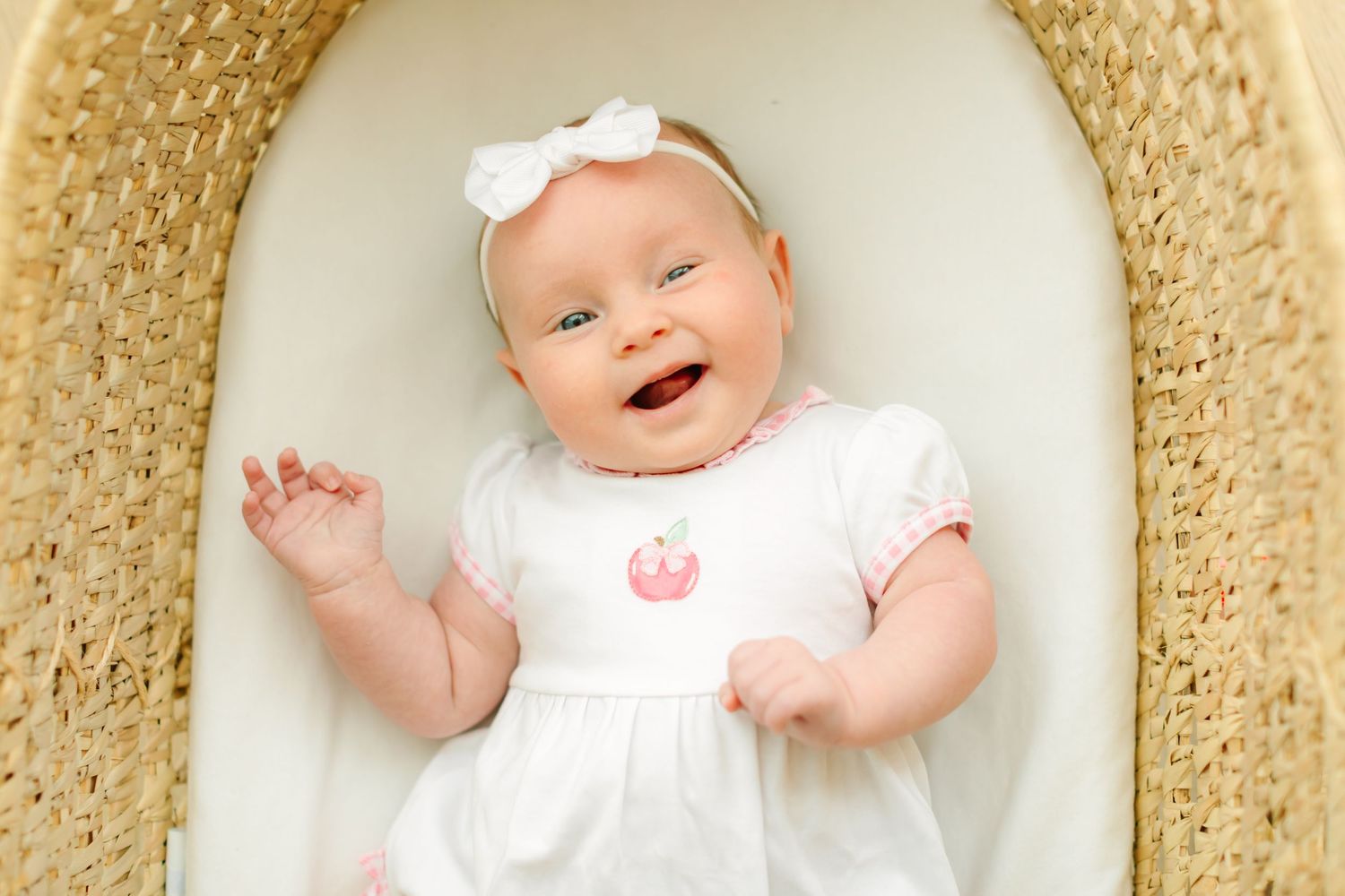A baby wearing white clothing smiles and waves while lying on a textured beige blanket.