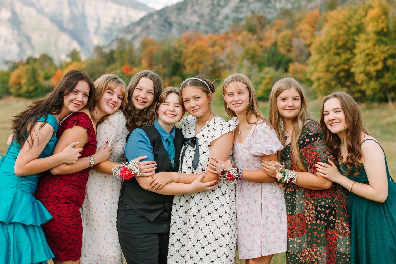 A group of friends in red and teal dresses pose together outdoors during autumn with colorful fall foliage in background.