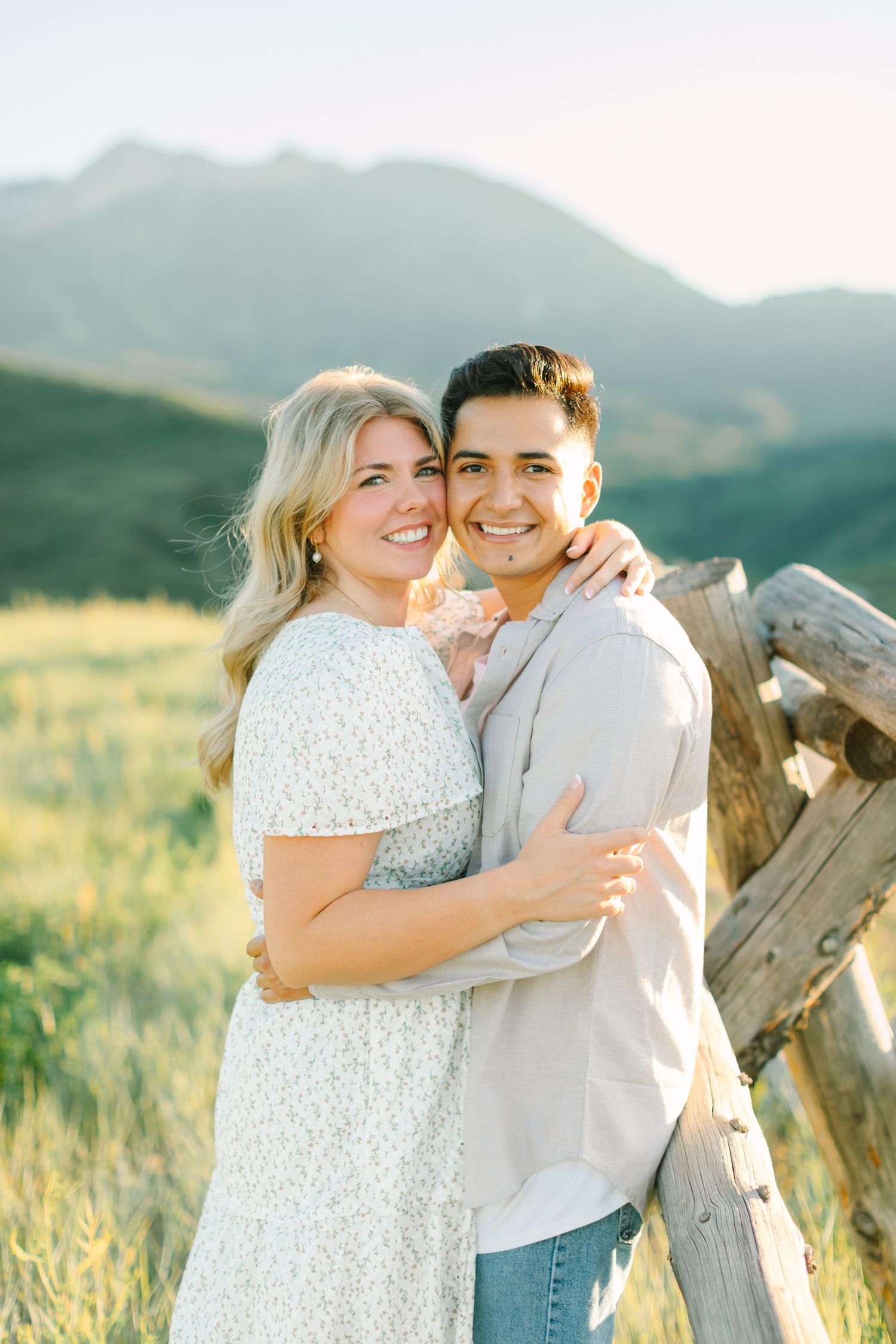 A couple embraces in a golden field at sunset with mountains and wooden fence in the background.