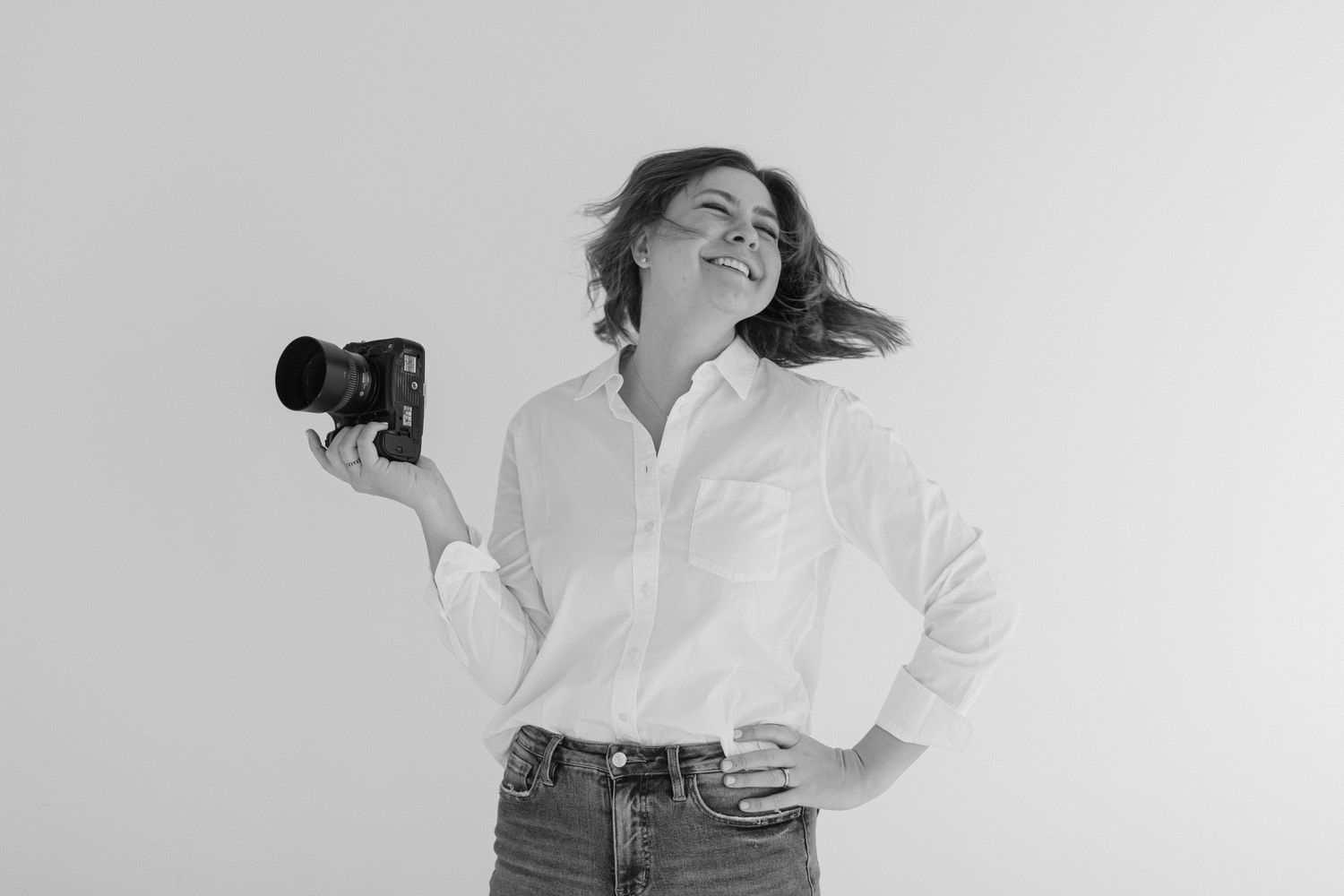 A photographer in a white button-down shirt and jeans holds a camera while posing against a white background.