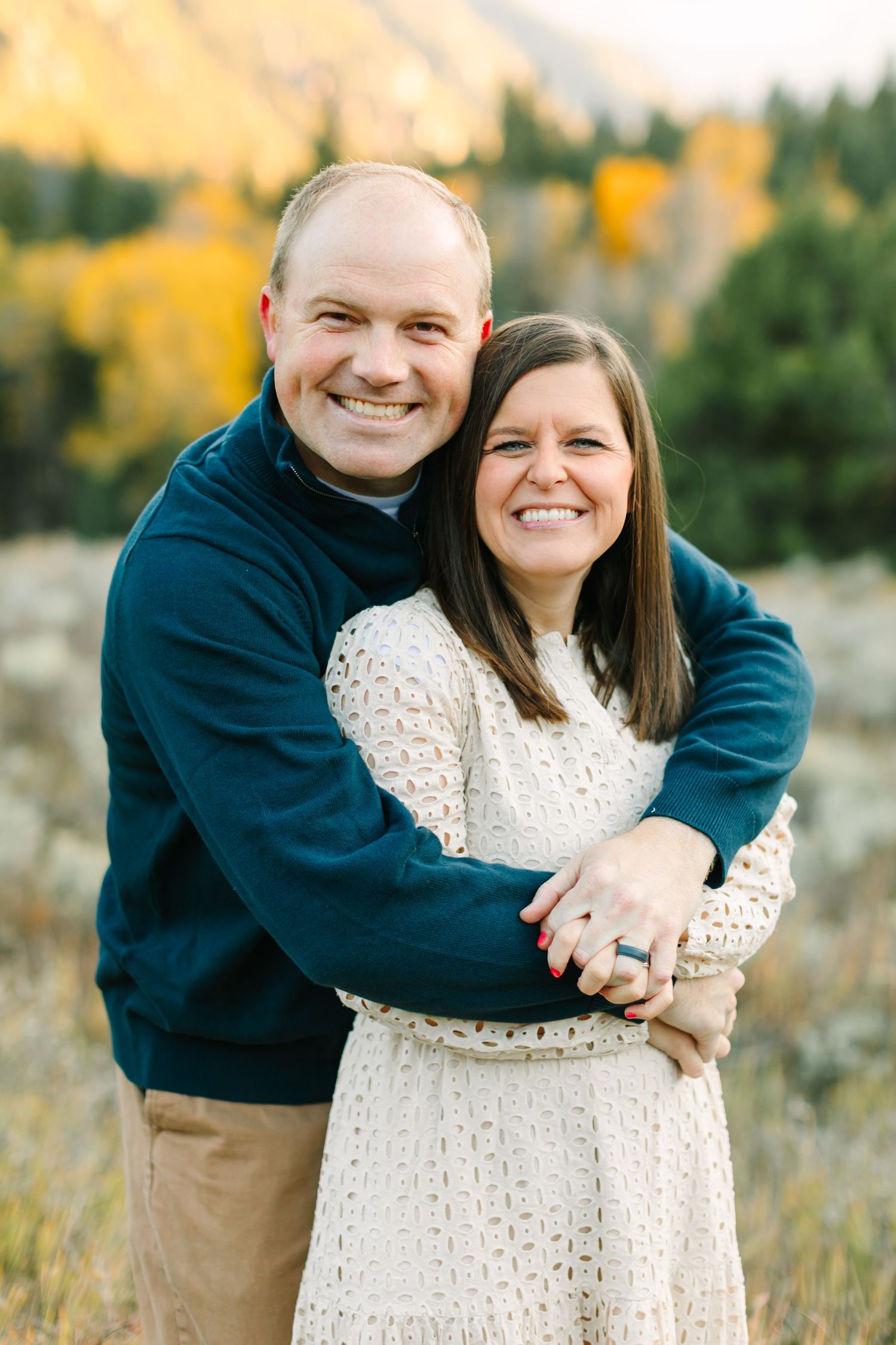 A couple embraces in a sunny field with autumn foliage in the background during an outdoor photo session.