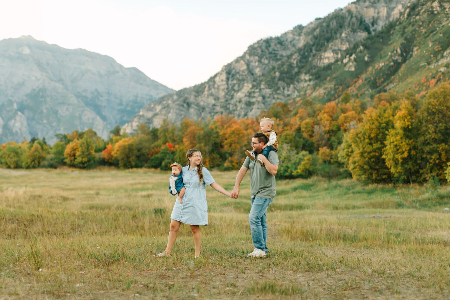 A couple walks through a scenic mountain meadow during autumn with golden grass and colorful fall foliage in the background.