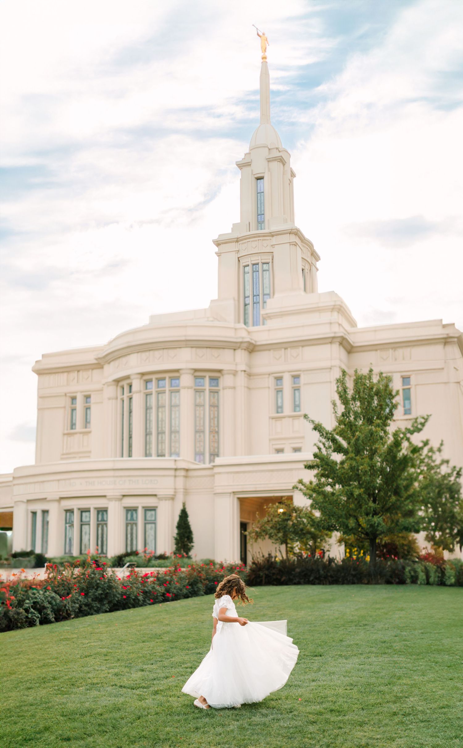 A bride in a flowing white dress twirls joyfully on the lawn in front of an elegant white temple building.