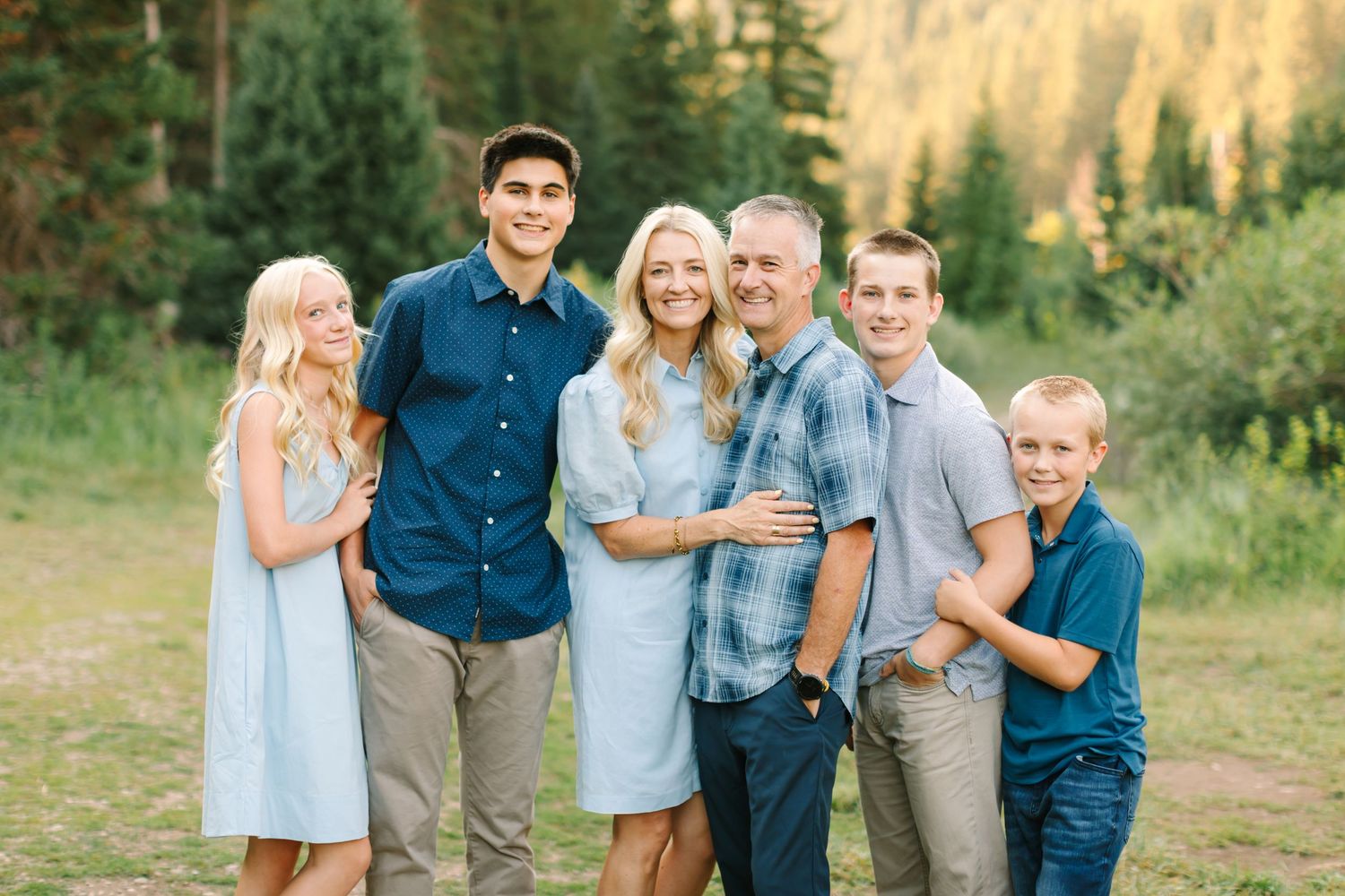 Happy family of six posing outdoors in a mountain forest setting, wearing coordinated blue and neutral outfits.