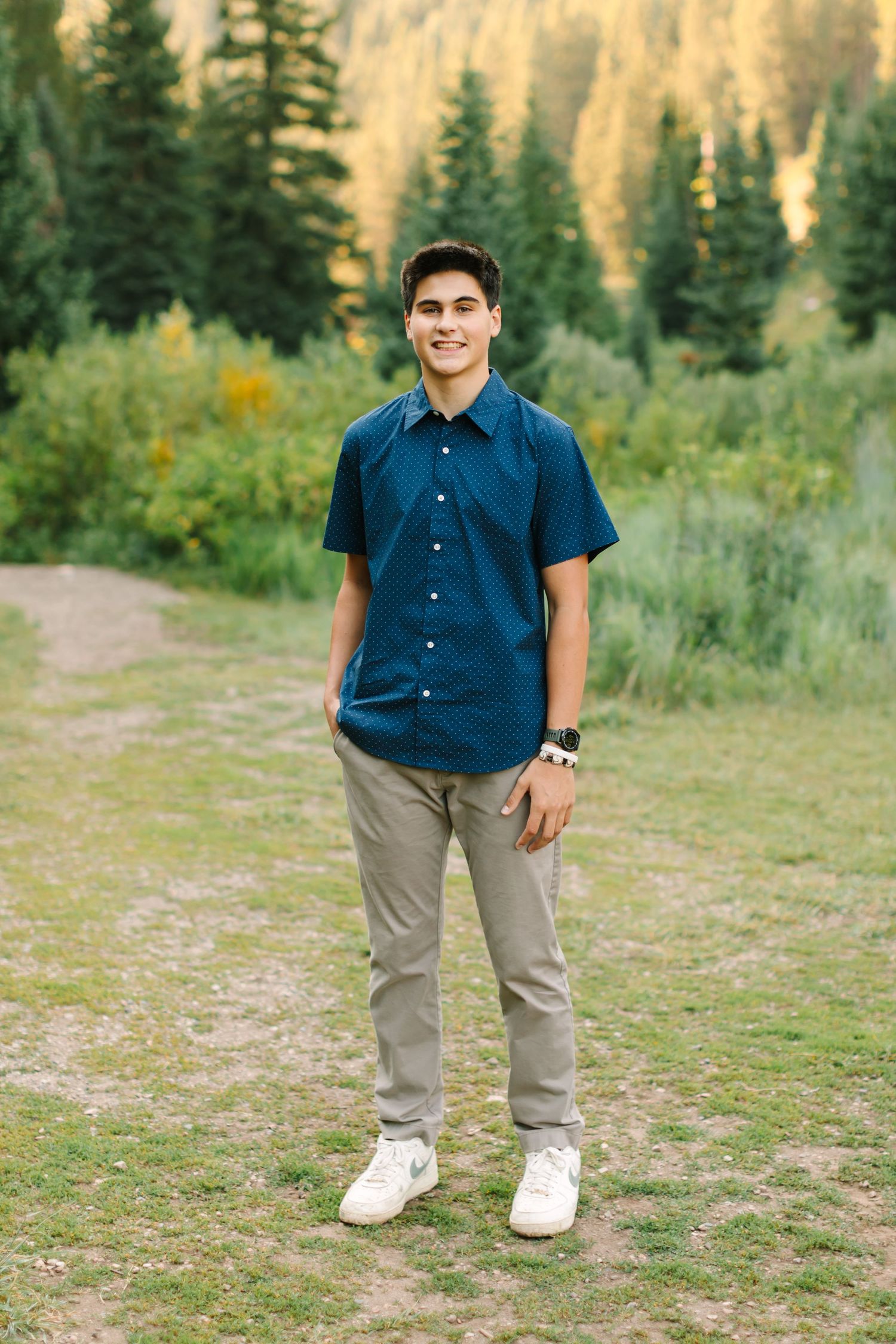 Young man in blue shirt and khaki pants smiling outdoors in a lush green forest setting.