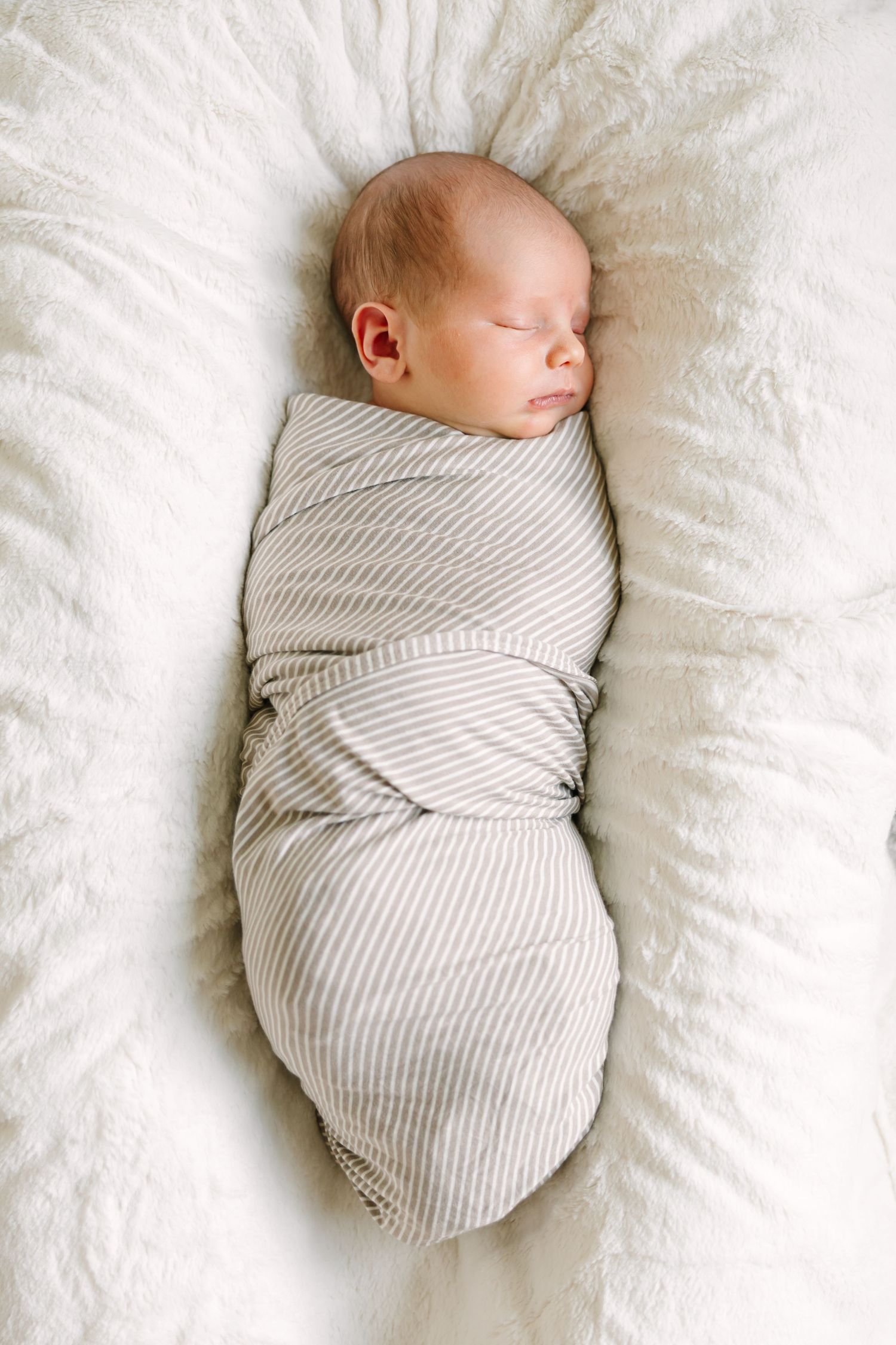 A sleeping newborn baby swaddled in a gray striped blanket lies peacefully on white bedding for his newborn session in Springville, Utah.