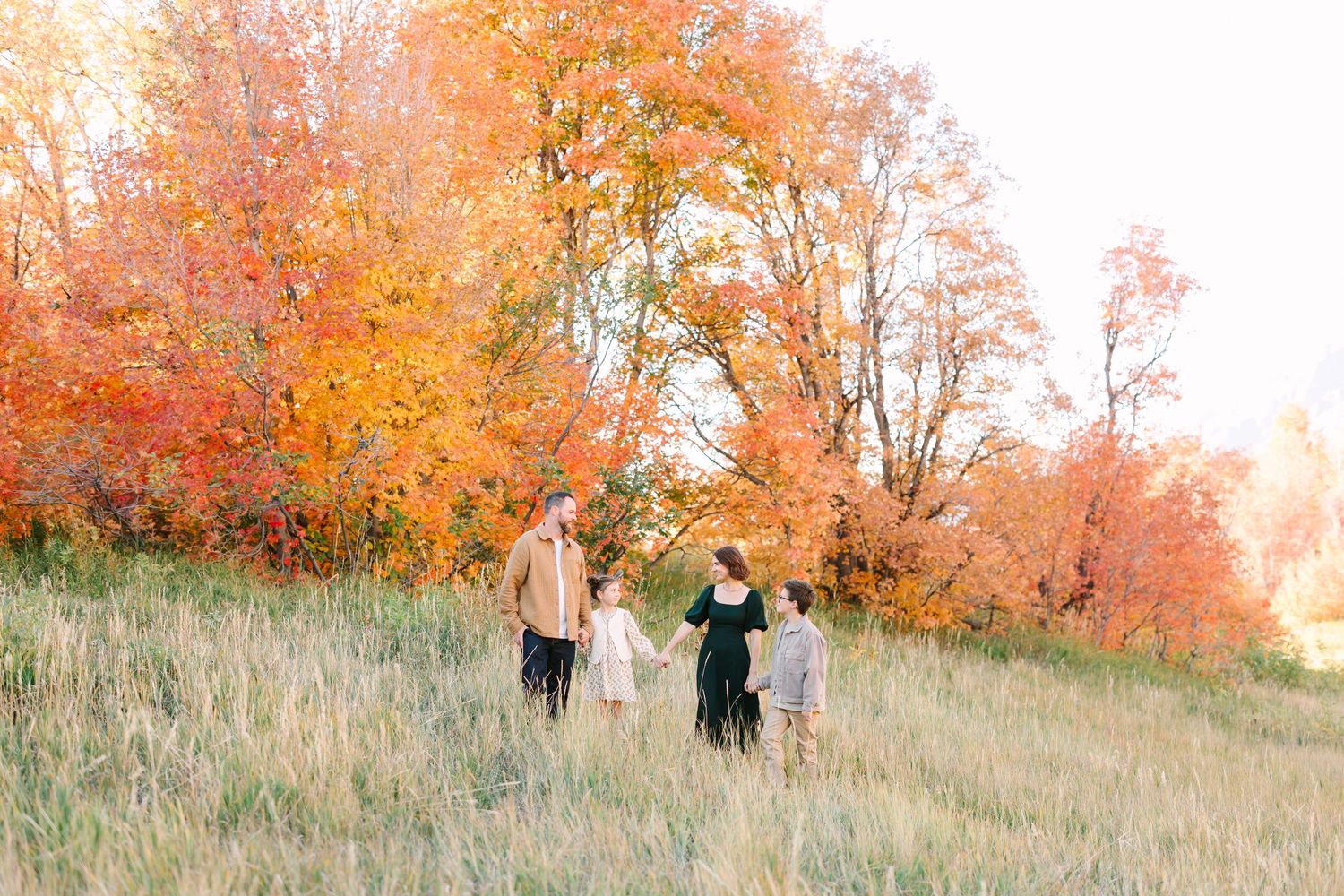 Three people walk together through a grassy hillside surrounded by vibrant autumn trees with orange and yellow foliage.