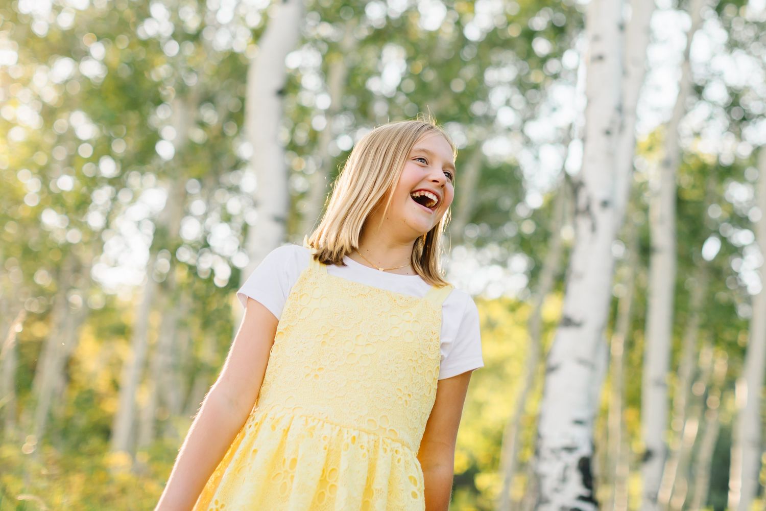 A person in a yellow dress twirls joyfully among tall birch trees in a sunlit forest setting.