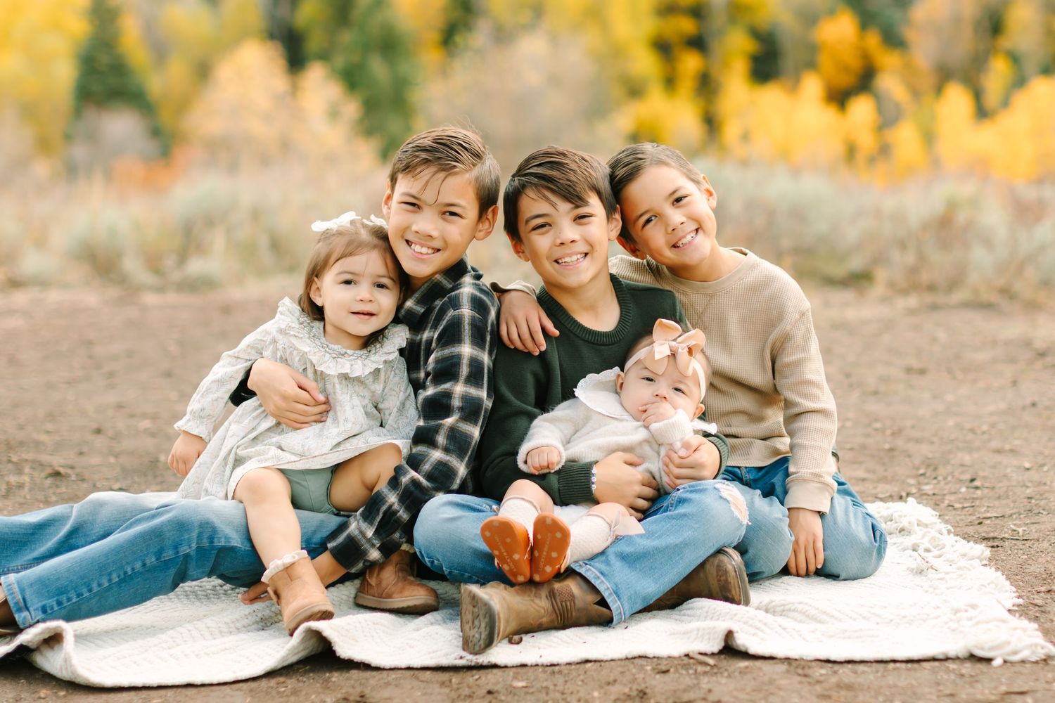 Family sitting together on white blanket outdoors with autumn foliage and golden trees in background.