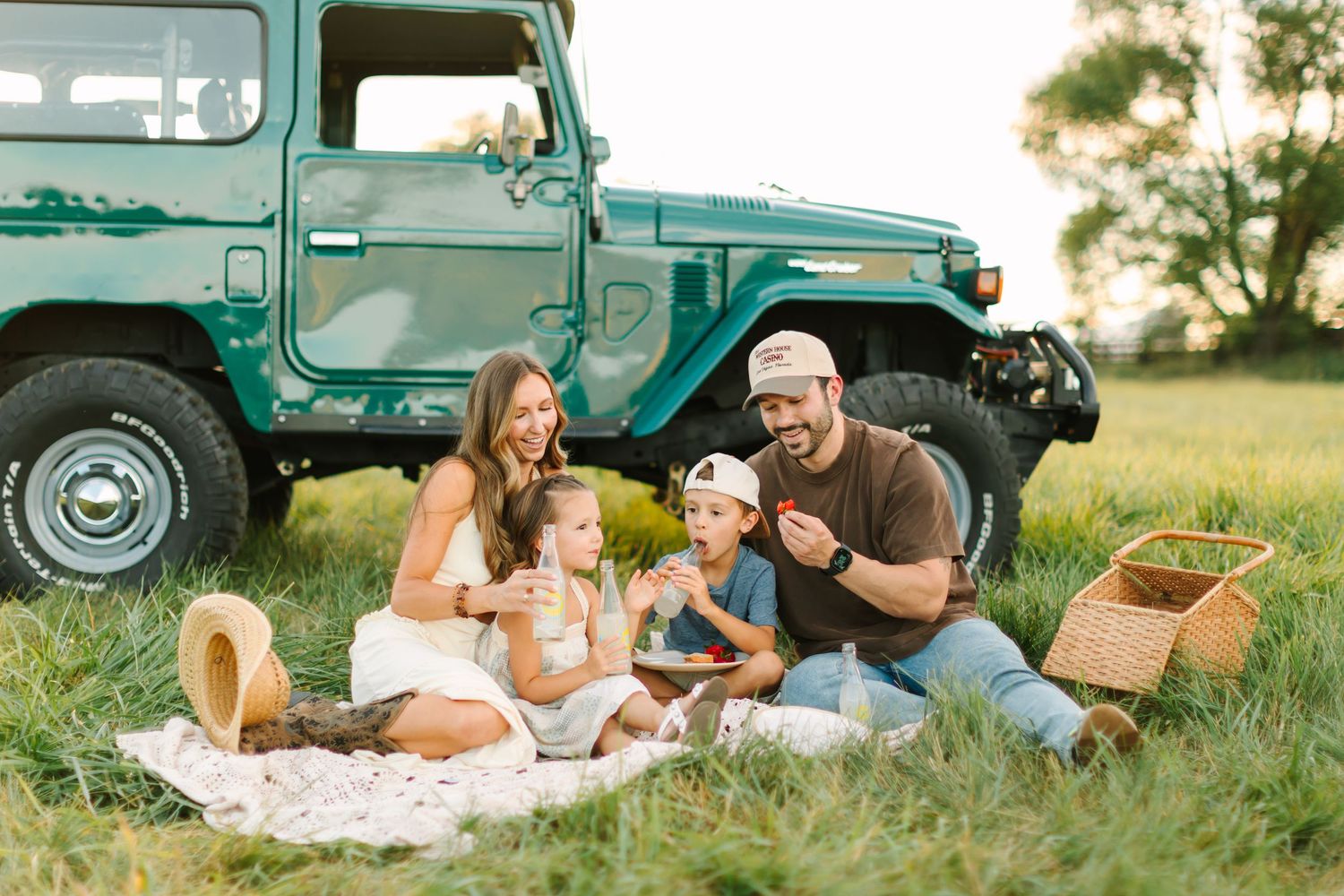 Couple having a romantic picnic date near a vintage turquoise truck in a golden summer field at sunset.