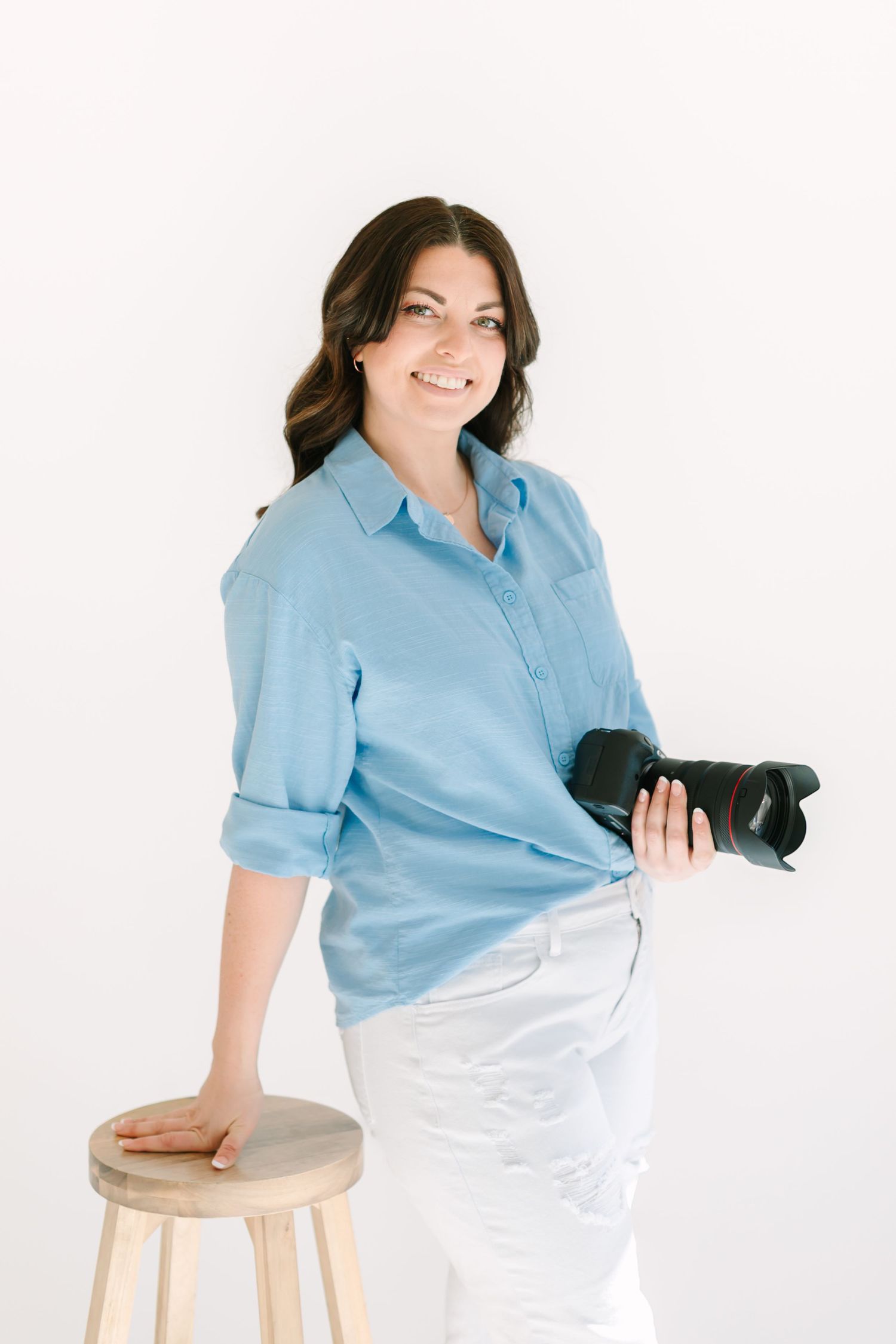 A photographer in blue shirt and white pants sits casually on a wooden stool in a bright studio.