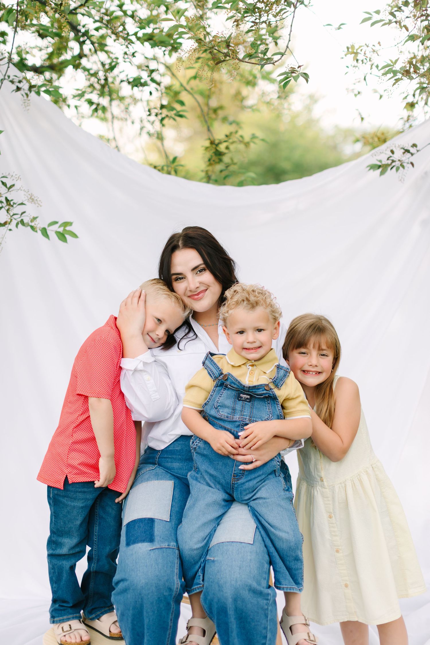 A mother sits outdoors holding her toddler while two children embrace her, posing in front of a white fabric backdrop.