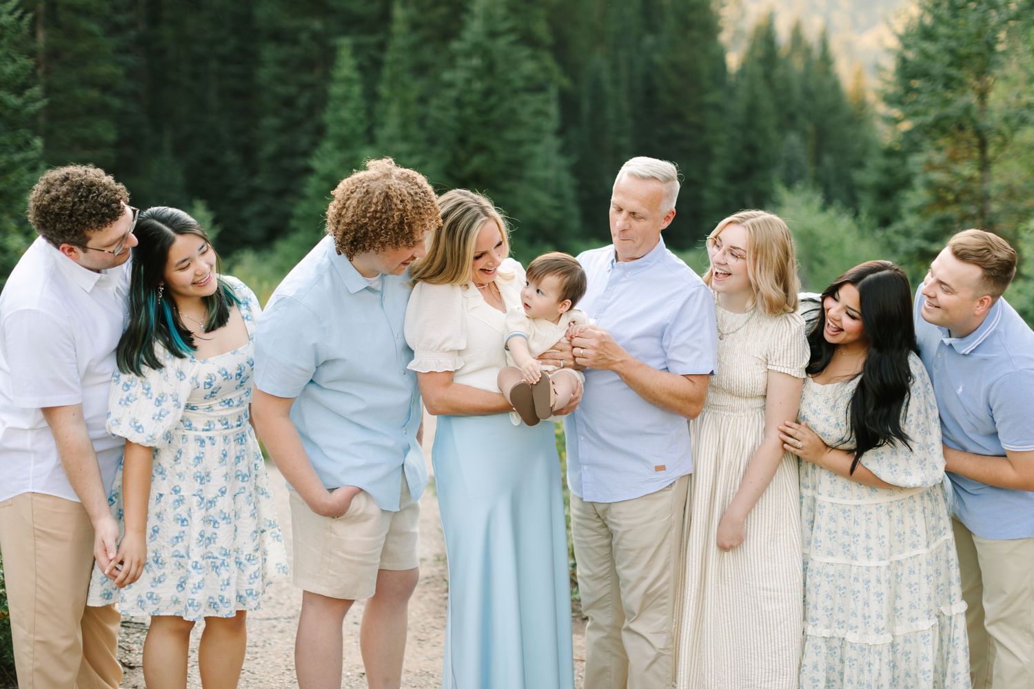 Large family group photo outdoors with pine trees, posing together in light neutral tones, sharing hugs and kisses in Utah.