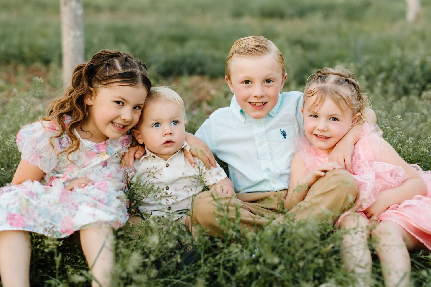 Four young children smile together outdoors, dressed in pastel spring outfits, sitting in lush green grass.