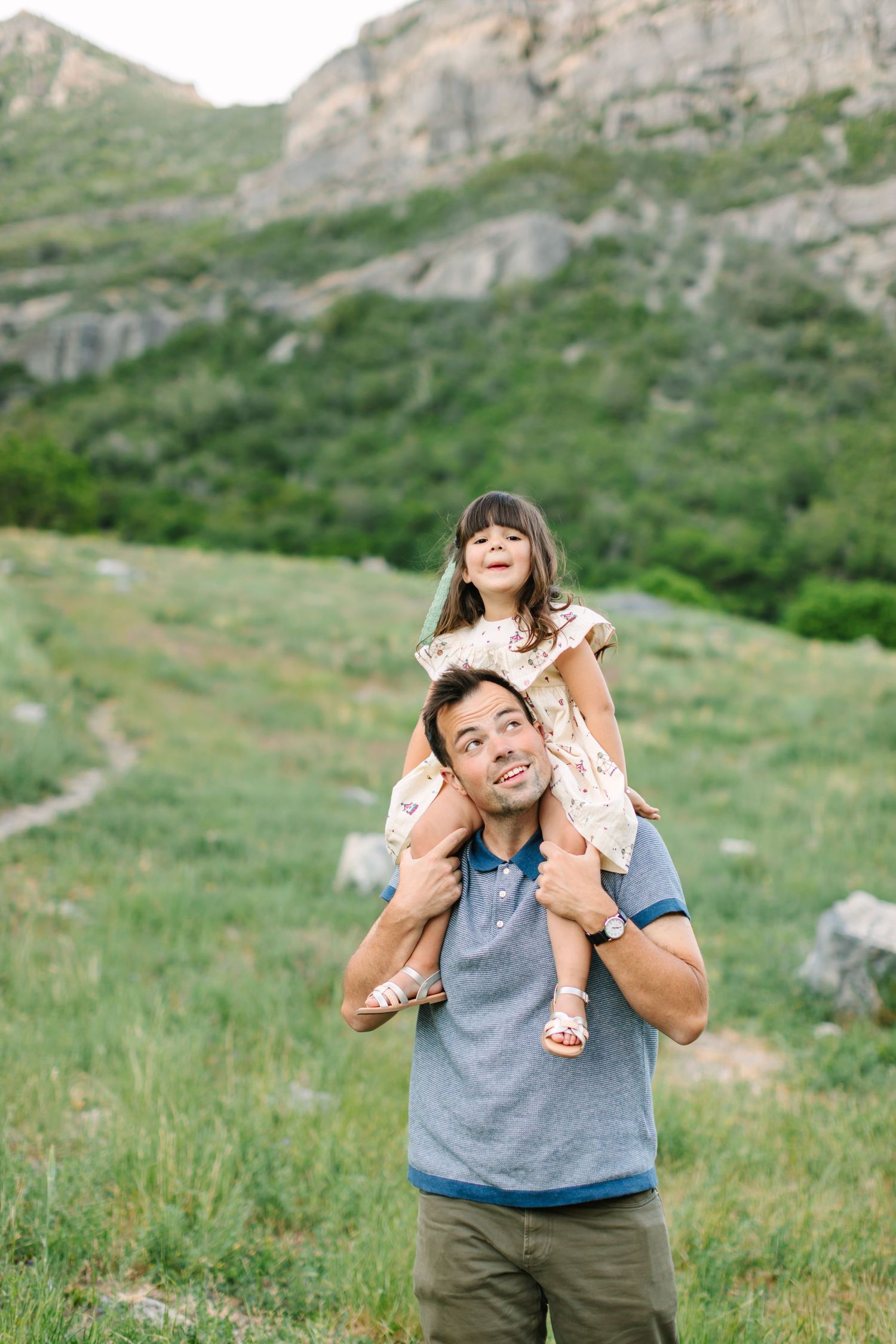 A happy father carries his laughing daughter on his shoulders in a lush green mountain meadow.