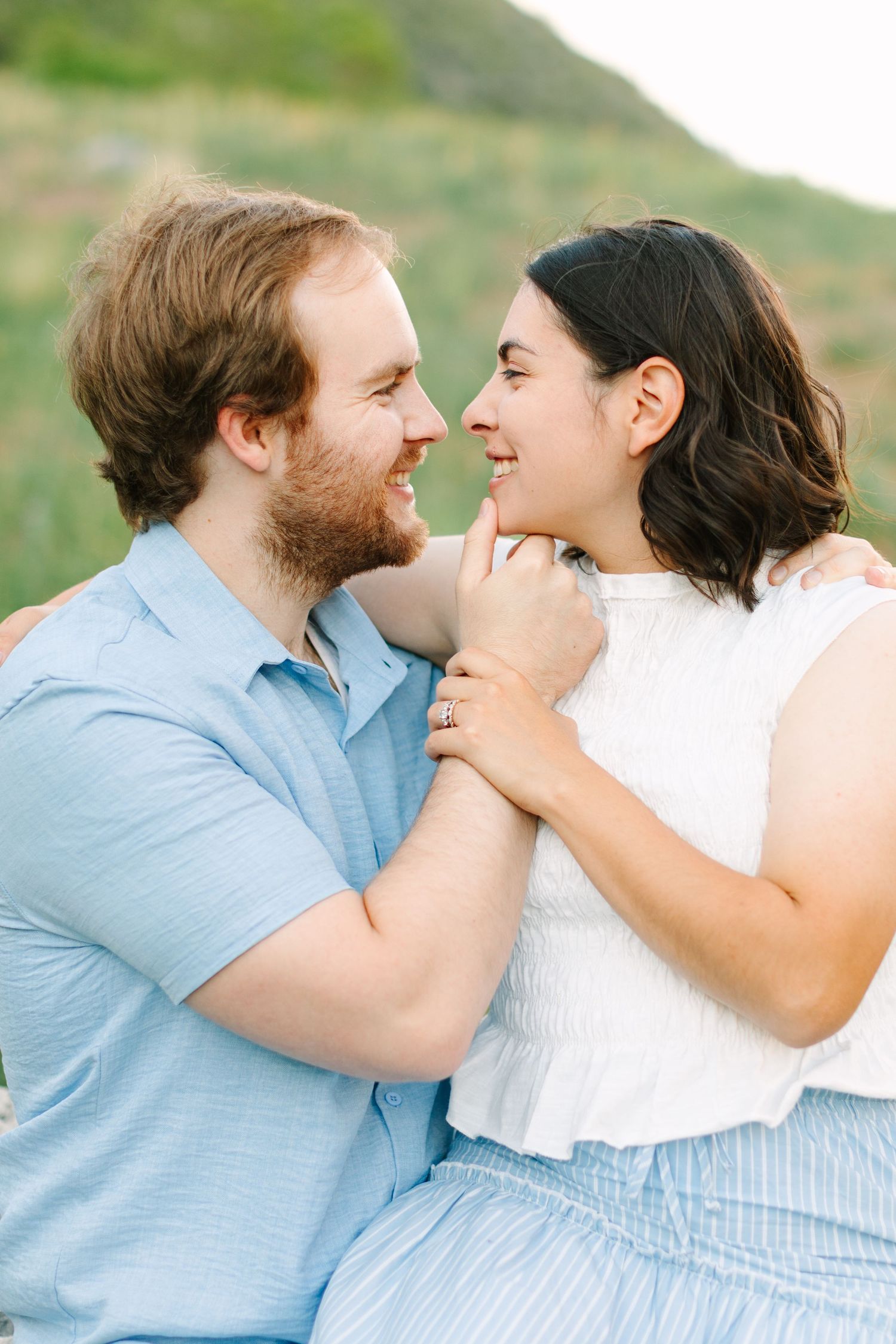 A smiling couple gazes at each other outdoors, the man in blue holding the woman's chin tenderly in a grassy setting.
