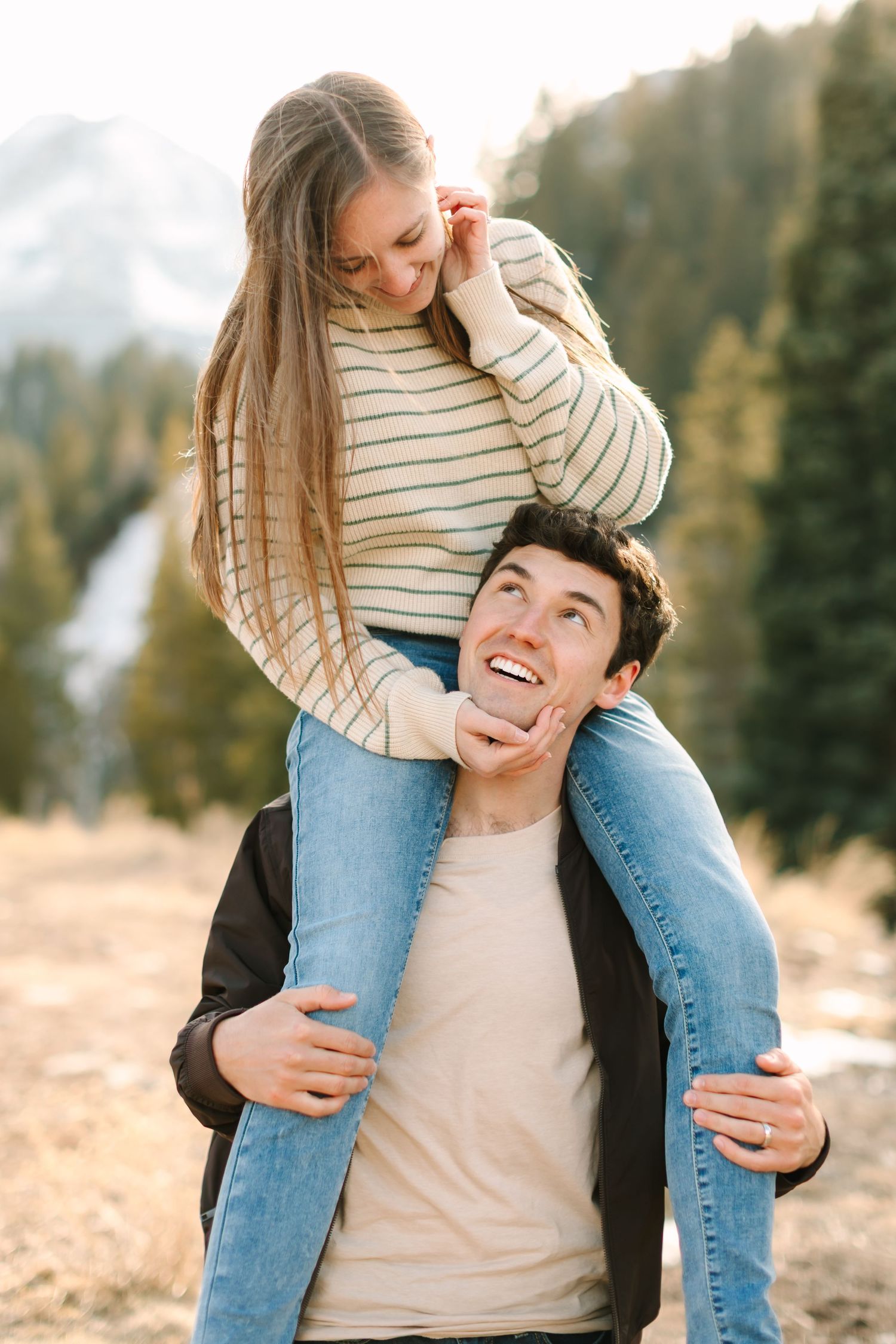 A joyful couple shares a playful moment outdoors with mountains and forest in the background.