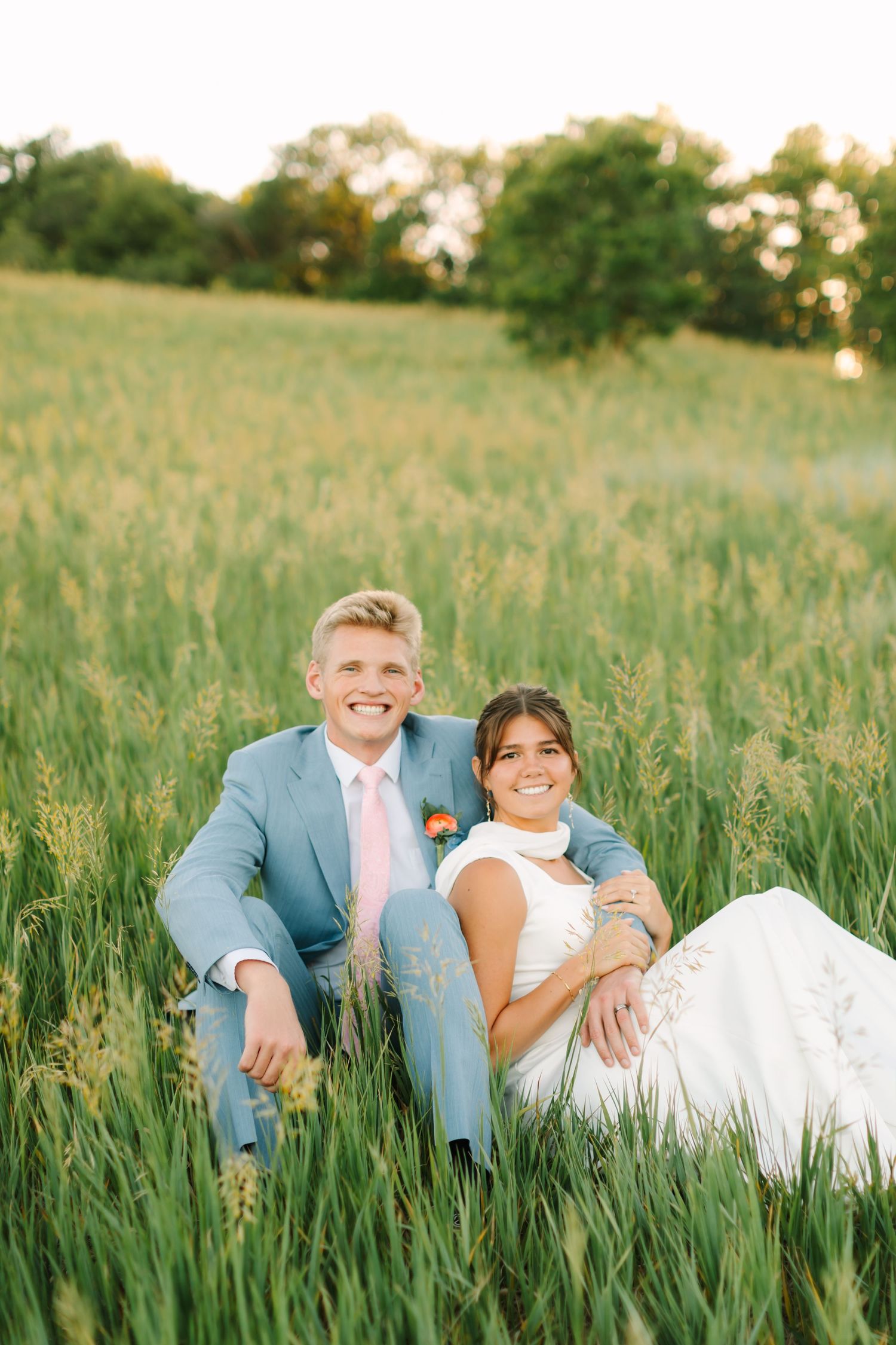 A smiling bride and groom sit together in a lush green field, wearing a white dress and light blue suit with pink tie.