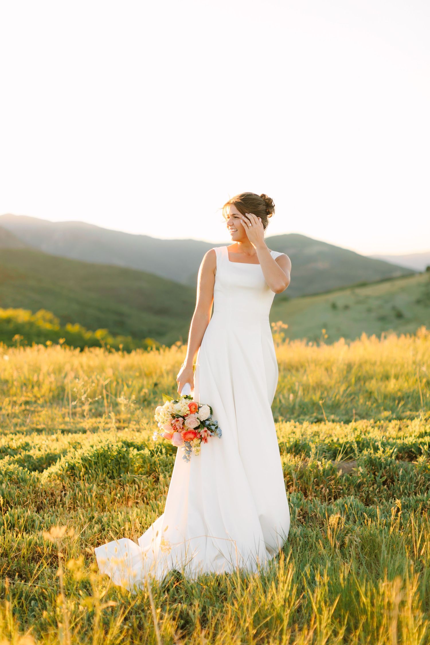 Bride in white gown holding colorful bouquet stands in golden meadow with mountain backdrop at sunset.