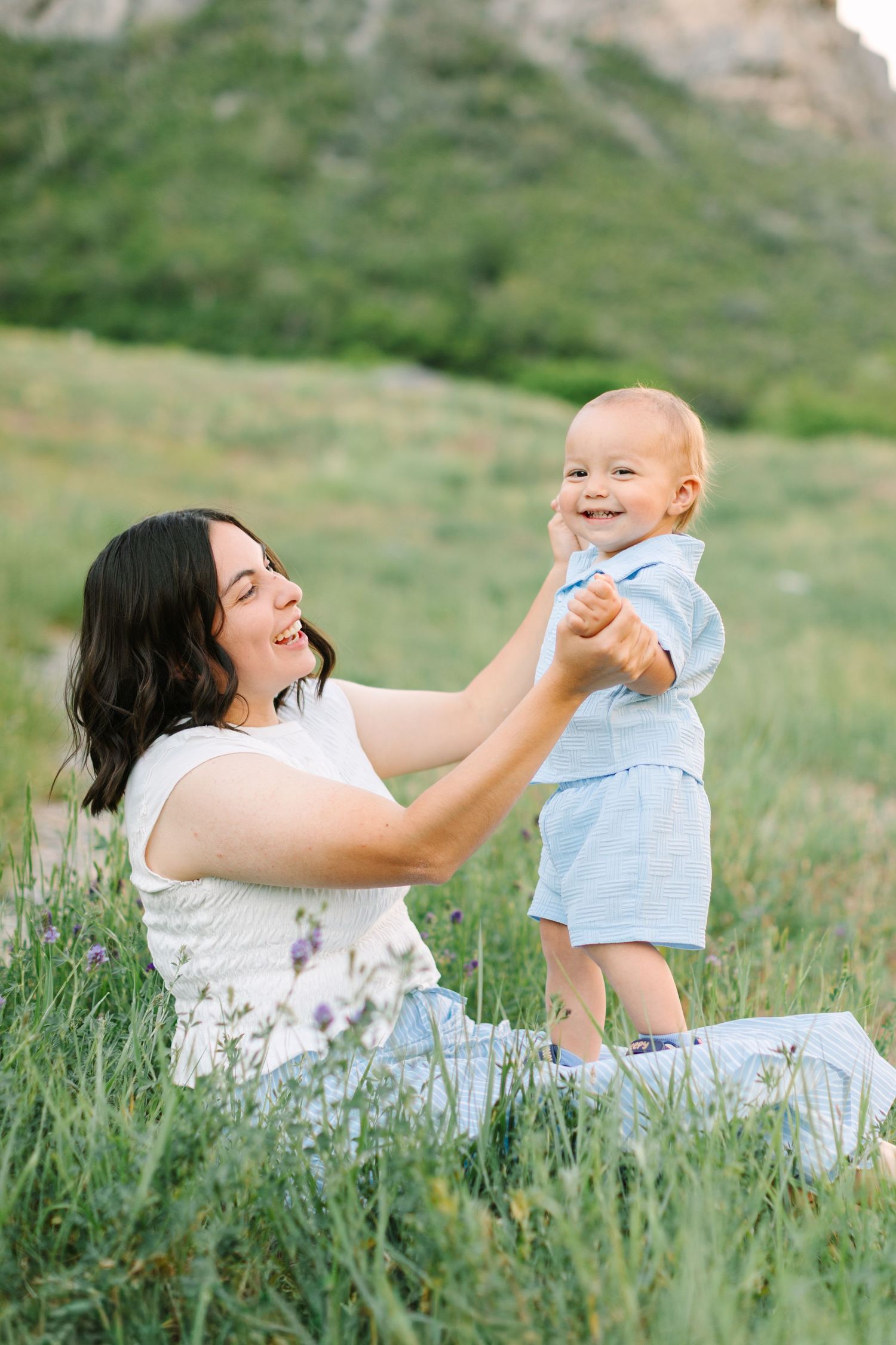 A smiling mother sits in a green meadow holding her happy baby boy in a light blue outfit on a sunny day.