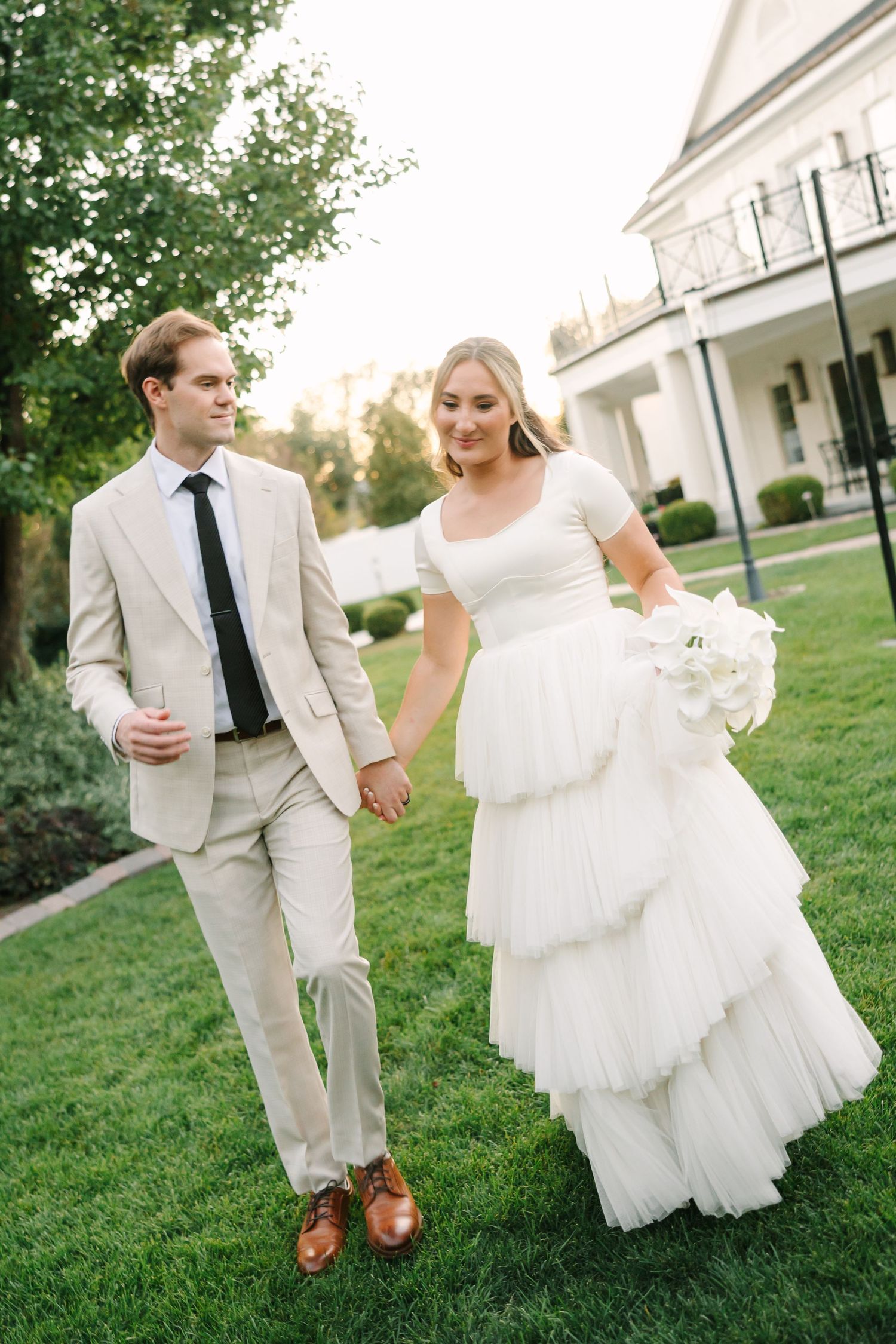 A bride and groom playfully run and dance together on a grassy lawn outside a white building during their wedding celebration.