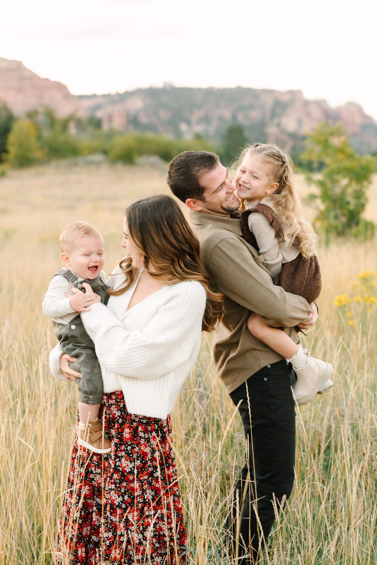 Family embraces and plays together in a golden field with mountain backdrop during sunset photo session.