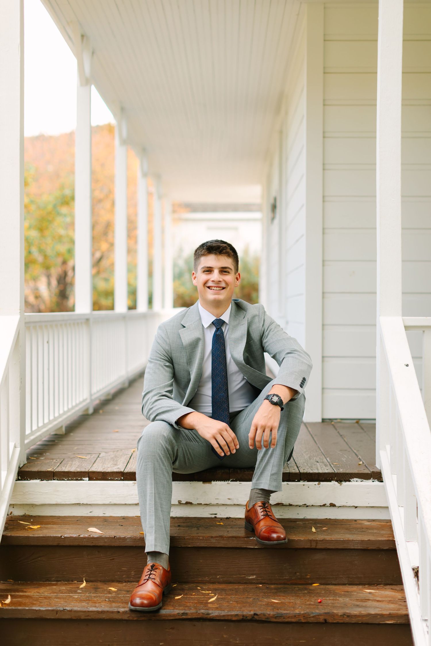 Two people in gray suits sit casually on a white porch with wooden floorboards.