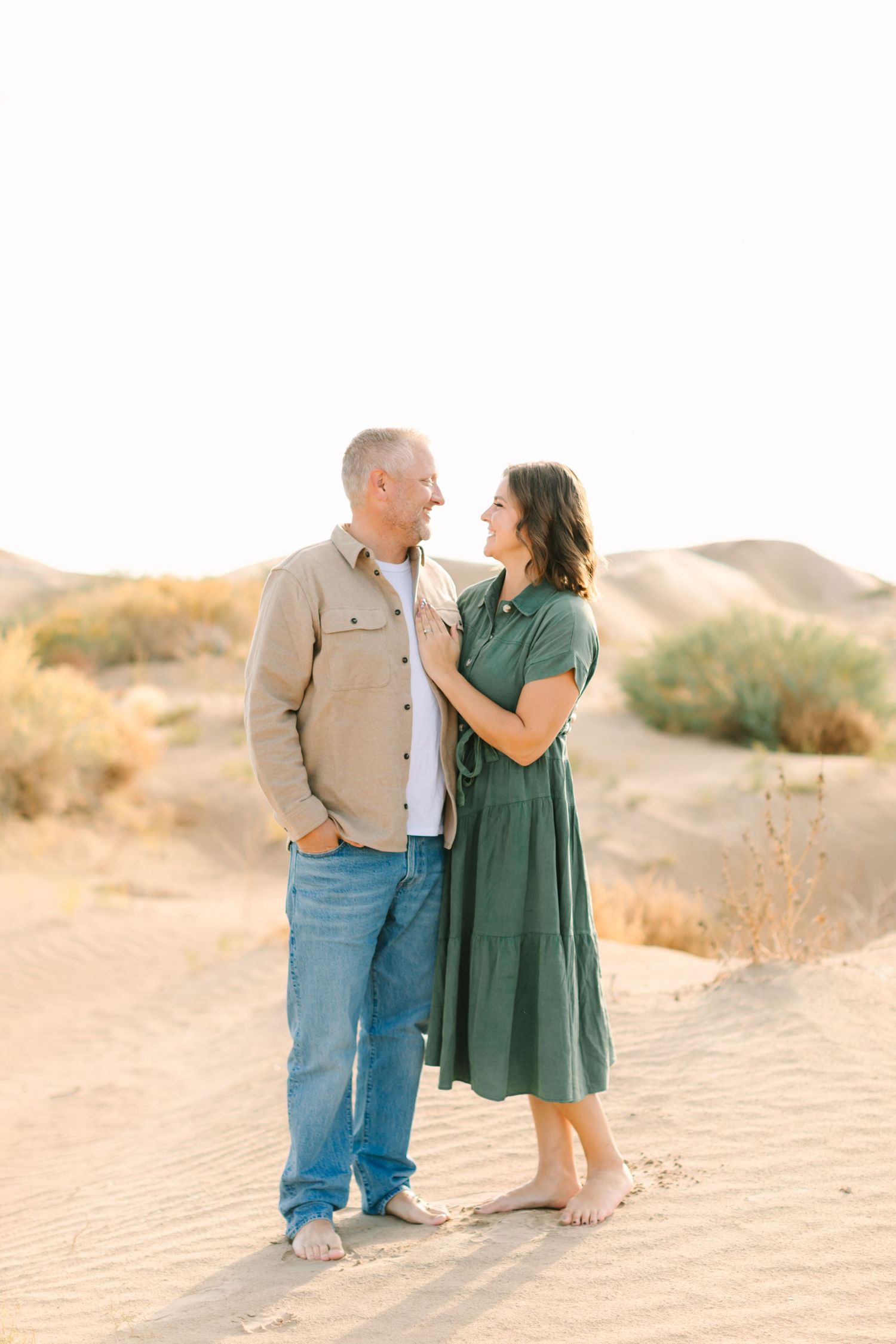 A couple shares a tender moment while standing together on sandy dunes during golden hour in a desert landscape.