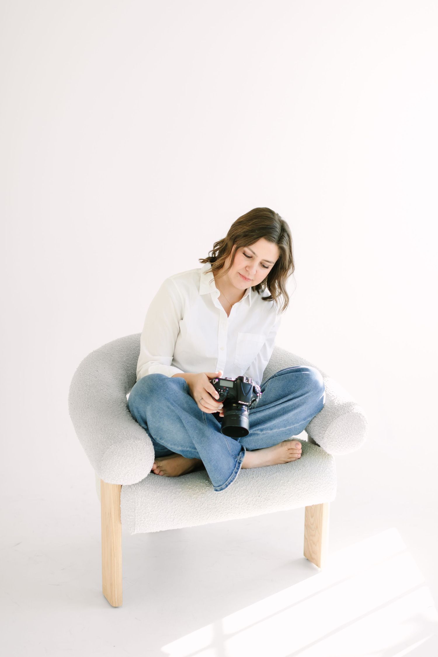Person in white shirt and jeans relaxes on white modern chair while holding camera in minimalist setting.