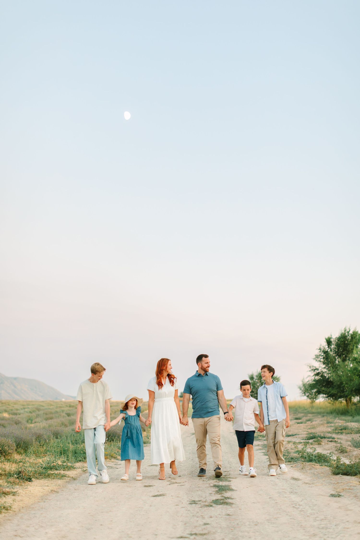 Family group walking together on a dirt path at sunset with mountains and moon visible in background.