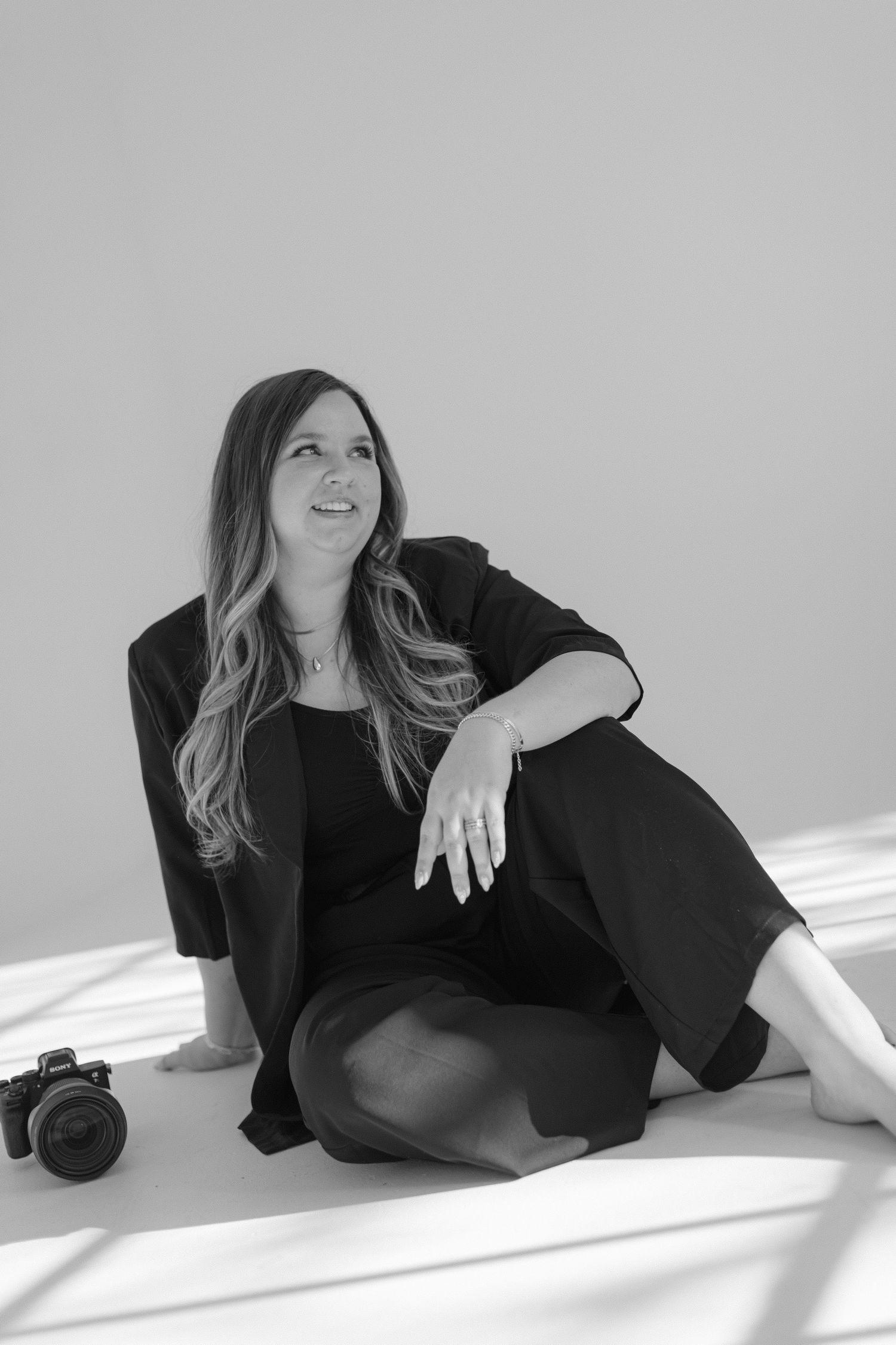 Black and white portrait of a person in dark clothing sitting on floor with a camera, looking up with natural window light.